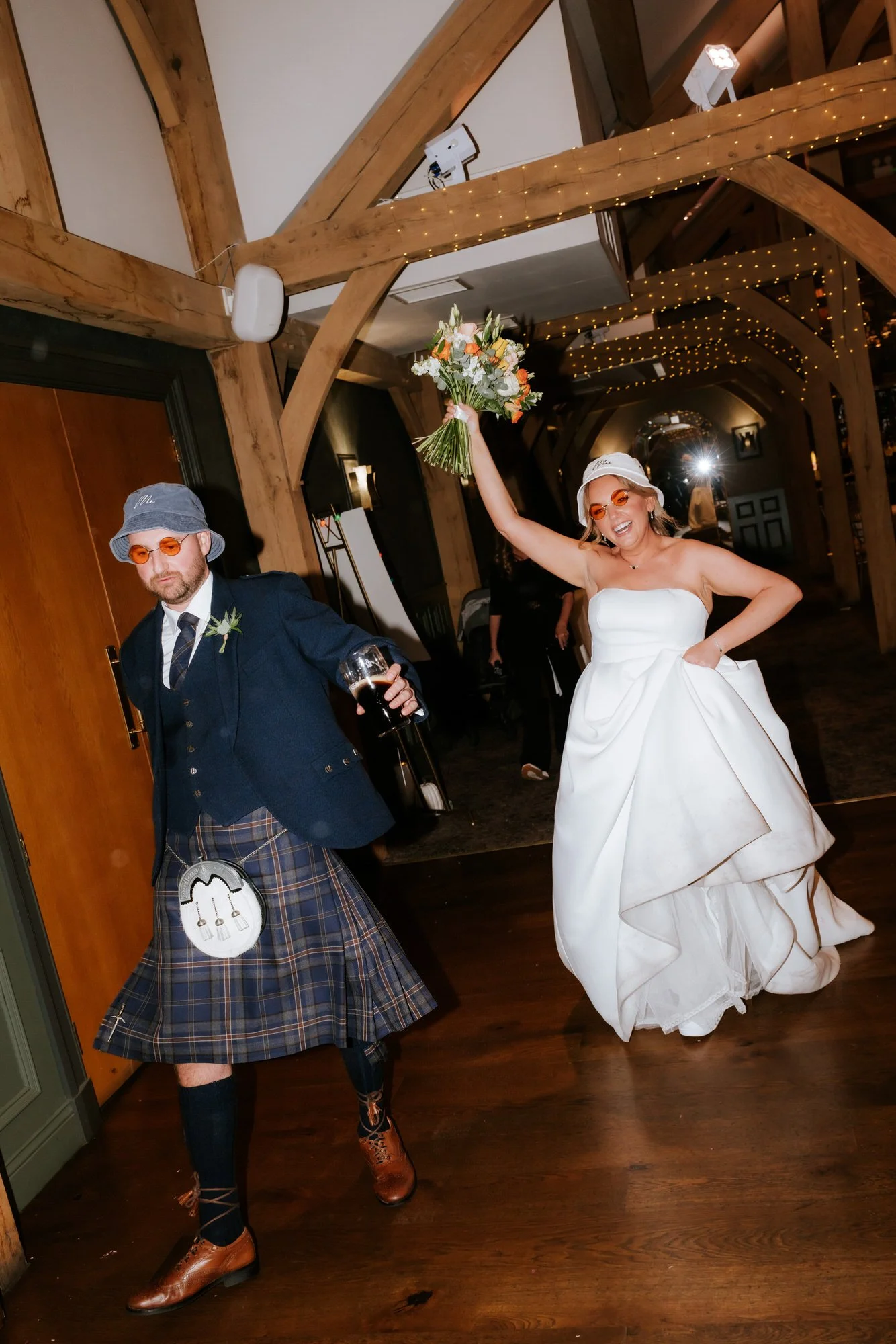 Wedding reception with a woman in a white wedding dress holding a bouquet and a man in traditional Scottish attire holding a drink, dancing in a warmly lit rustic hall.