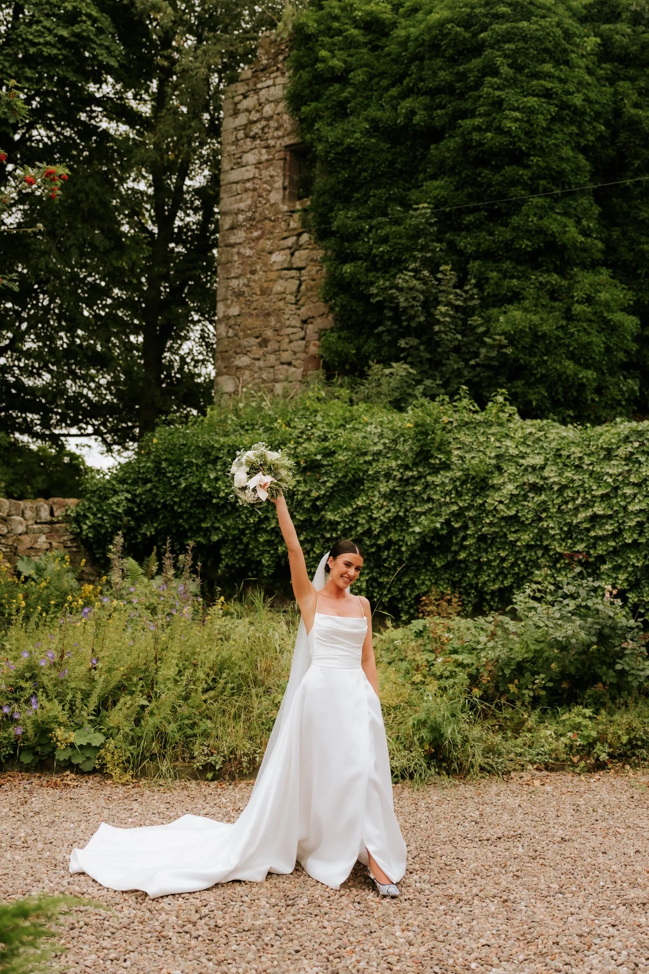 A joyful bride in a white wedding dress holding a bouquet up high, standing on a gravel path outdoors with lush green foliage and an old stone tower in the background.