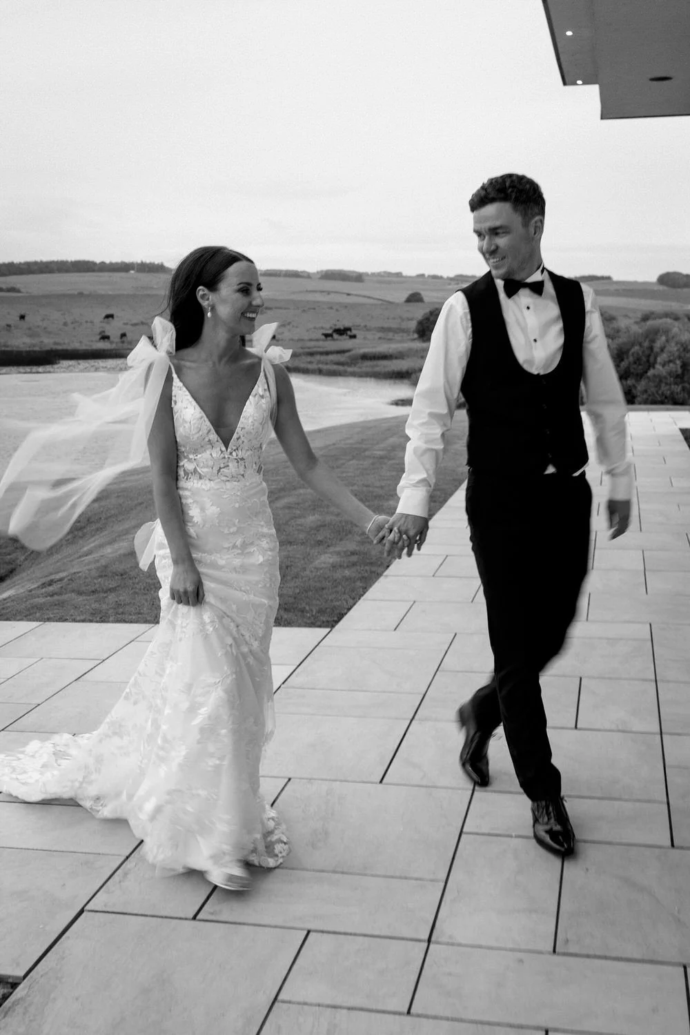 A bride and groom holding hands and walking outdoors, smiling at each other, during their wedding day.