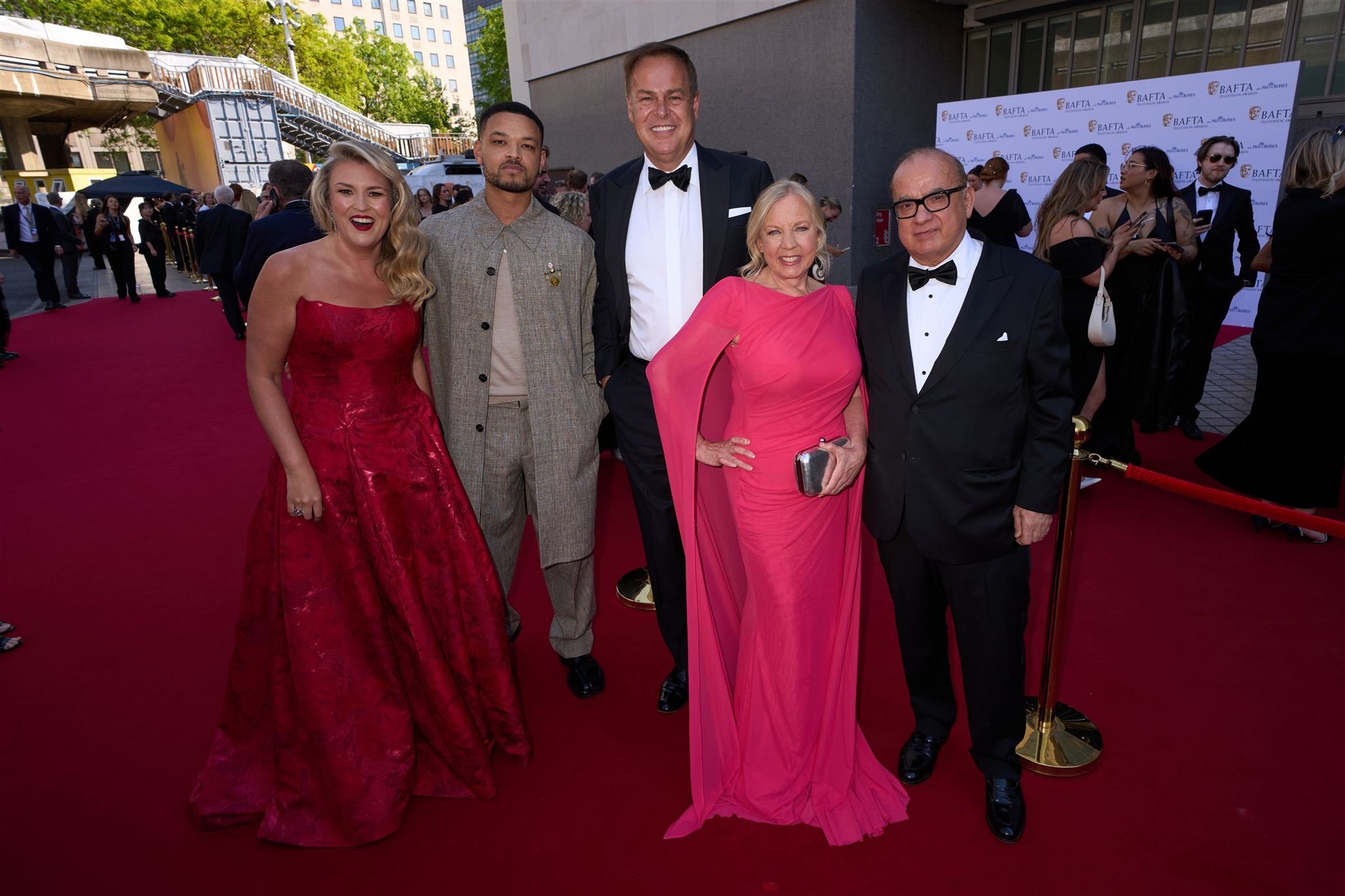 Group of five people standing on red carpet at a formal event, with a BAFTA backdrop in the background. They are dressed in elegant evening attire, with women in colorful gowns and men in tuxedos.