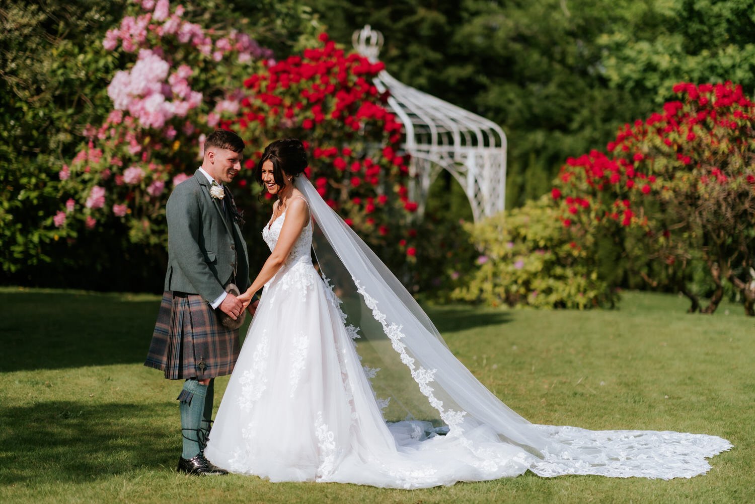 A bride and groom holding hands, smiling, outdoors in a garden with vibrant flowers and greenery, under a white gazebo.