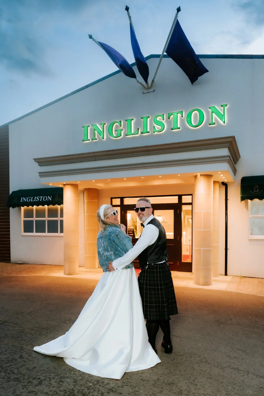 A couple dressed in wedding attire standing in front of ING_LISTON store, smiling and holding each other, with a cloudy sky overhead.