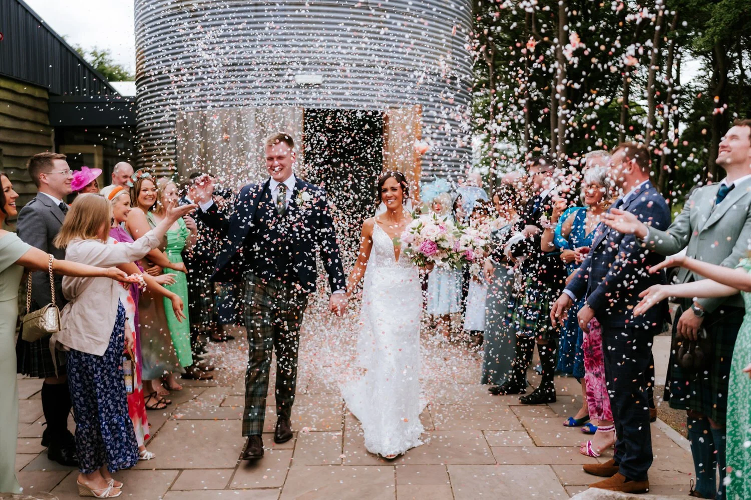 Bride and groom walking hand in hand through a crowd of friends and family, celebrating with confetti outside a modern building, during their wedding.
