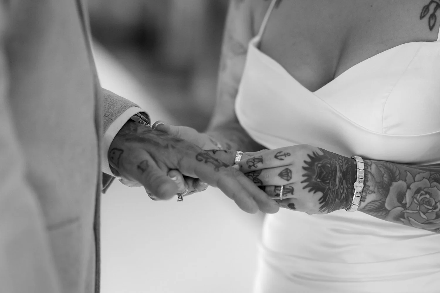 Close-up black and white photo of two tattoosed hands clasped together, one wearing rings and bracelets, the other in a long-sleeve shirt and a white dress.