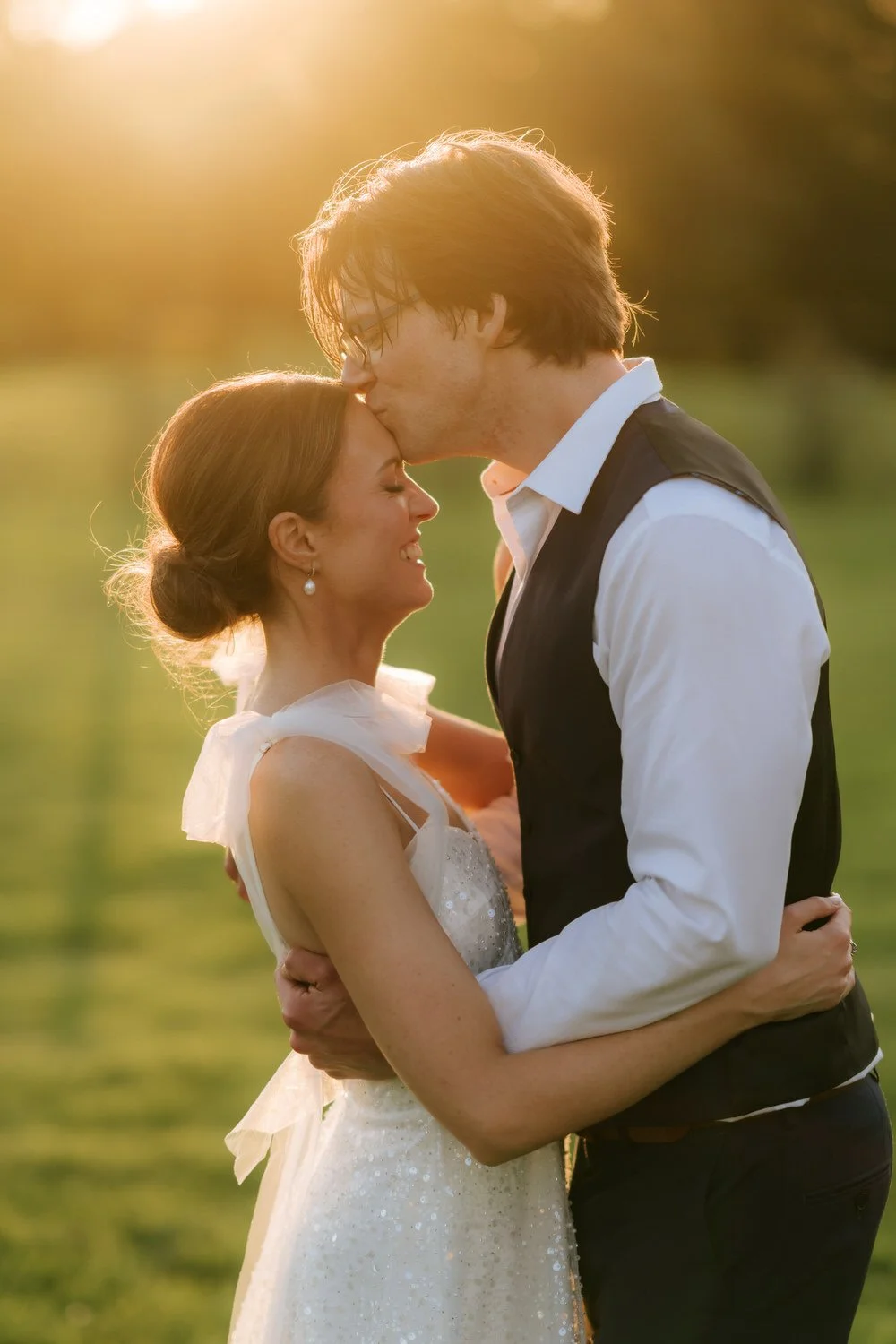 A bride and groom embrace outdoors in warm, golden sunlight. The groom kisses the bride's forehead as they hold each other close, with a blurred background of greenery.