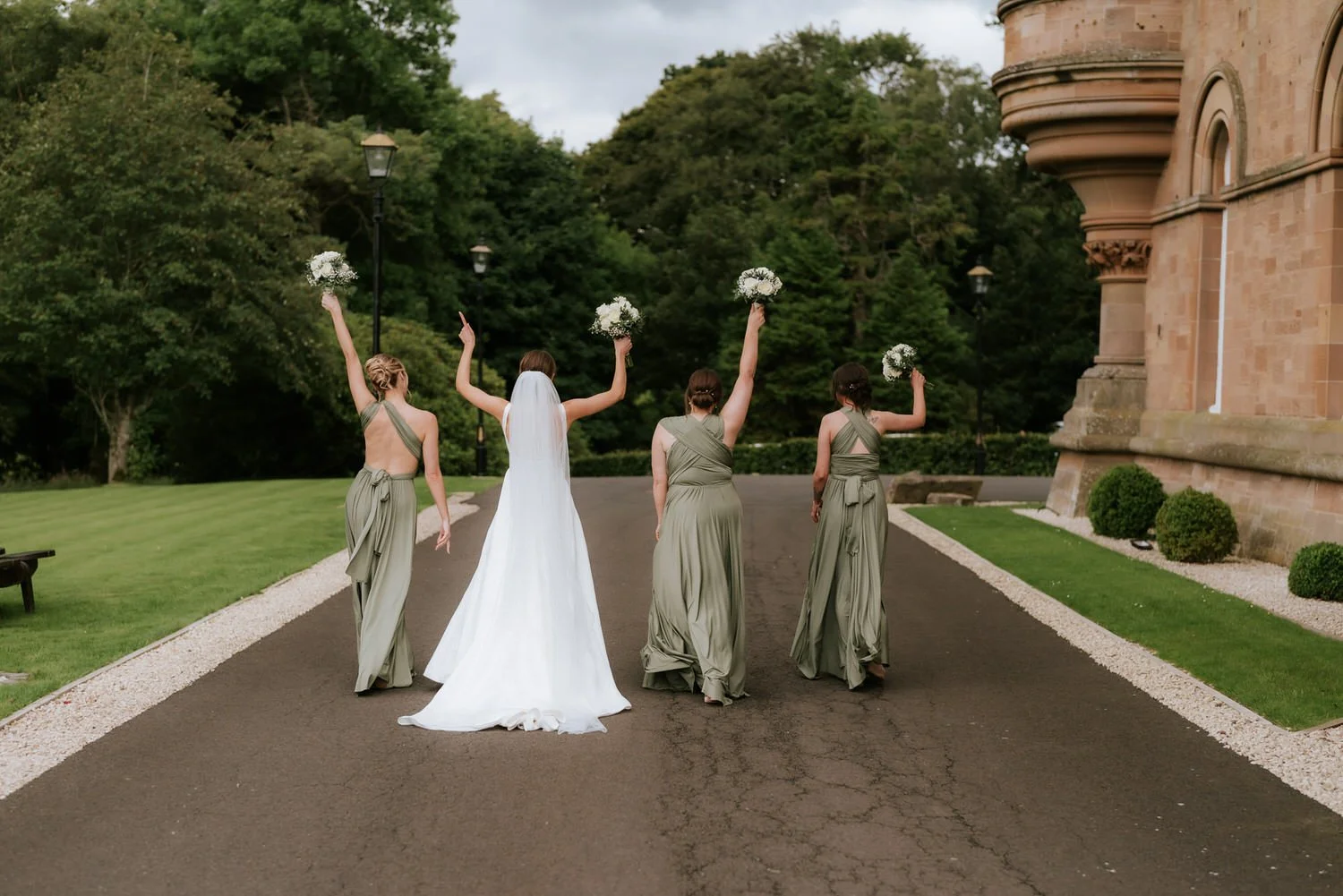Bride and four bridesmaids walking on a paved path outside, holding bouquets, near a brick building and lush green trees.