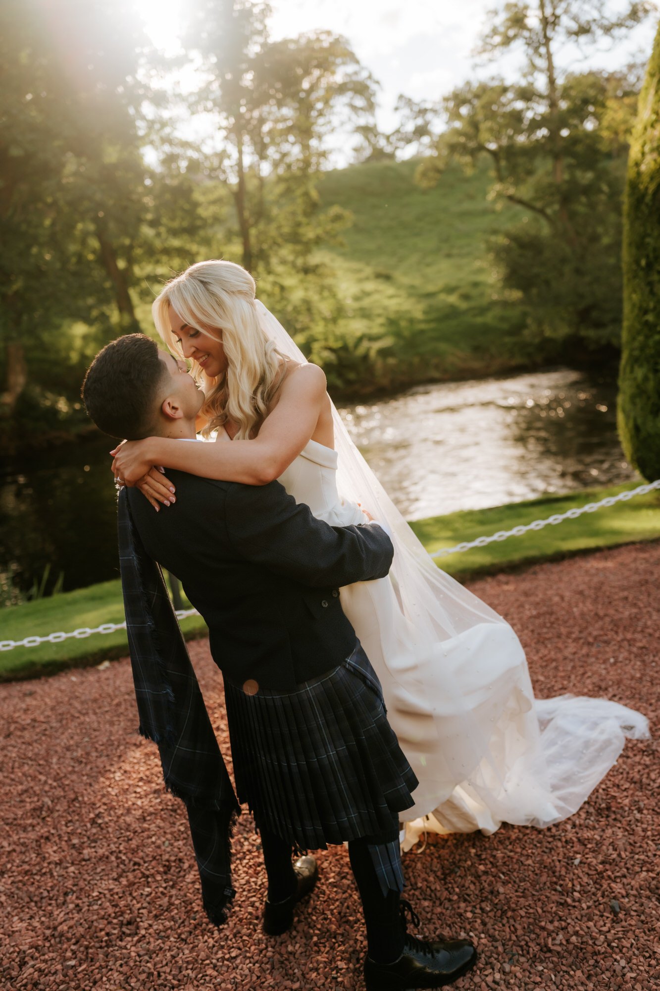A woman in a white wedding dress is lifting a young man in formal attire. They are outdoors near a river, with sunlight filtering through trees in the background.