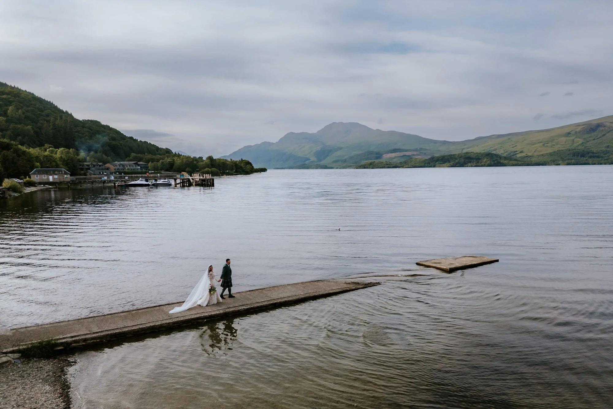 A bride and groom walk on a concrete pier by a lake with mountains in the background. The bride wears a white wedding dress and veil, and the groom wears a dark suit and kilt. Overcast sky.