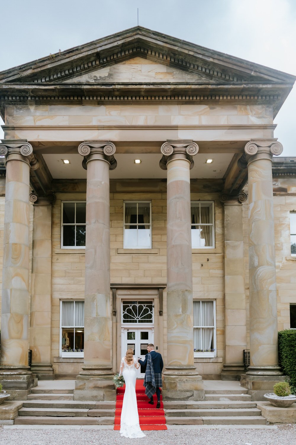 A bride and groom walking up a red carpeted staircase in front of a historic building with large stone columns and an ornate doorway.