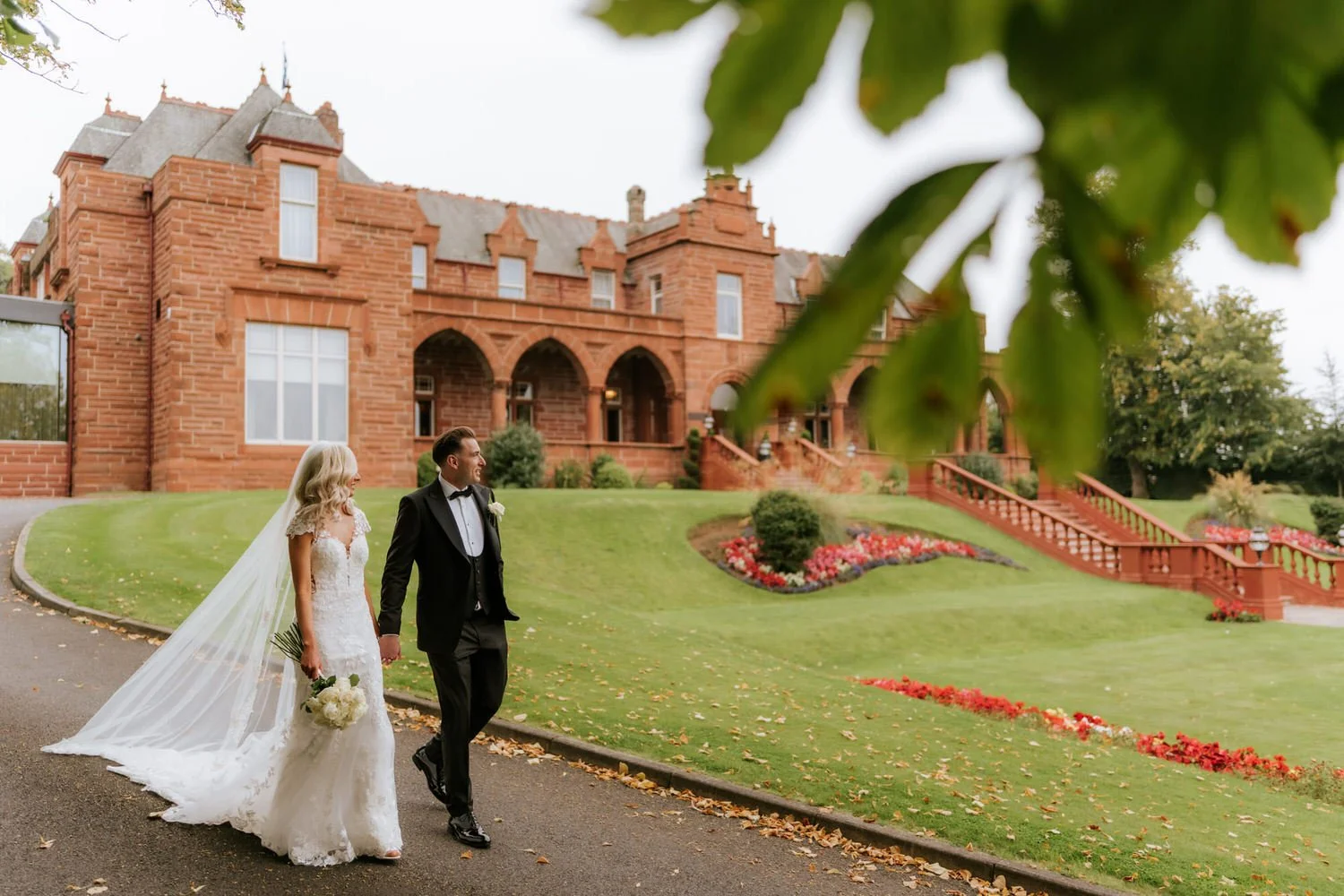 Bride and groom walking hand in hand outside a large brick mansion on a cloudy day. The bride is wearing a white wedding gown with a long veil, holding a bouquet of white flowers. The groom is dressed in a black tuxedo. The mansion has a grand stairc