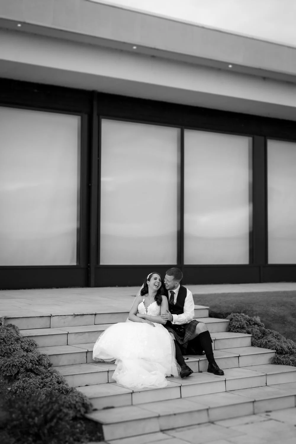 A bride and groom sitting on outdoor steps, smiling and laughing together, with large windows in the background.