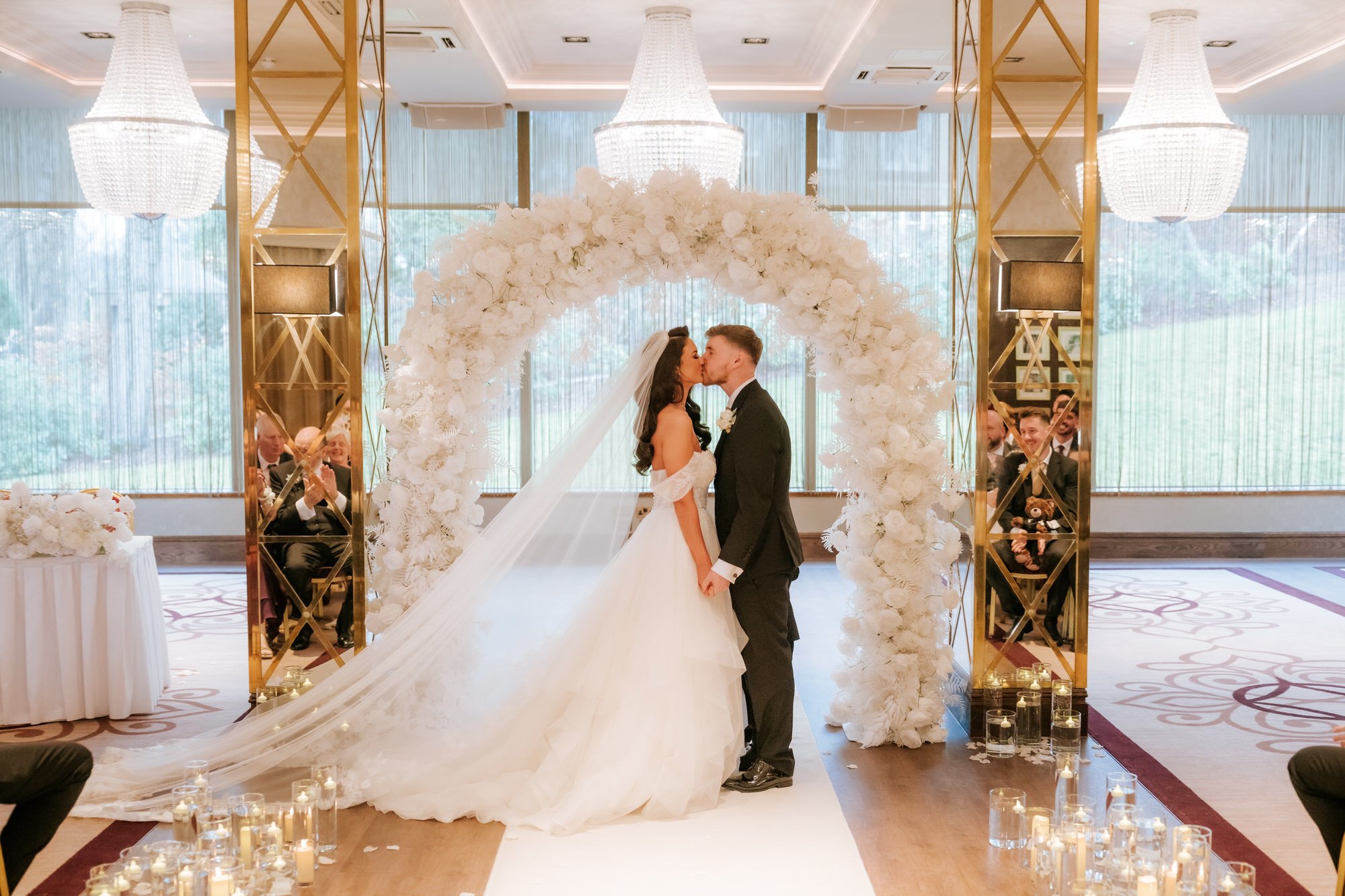 A bride and groom sharing a kiss during their wedding ceremony in an elegant, decorated venue with a large white floral arch, chandeliers, and guests seated on either side.
