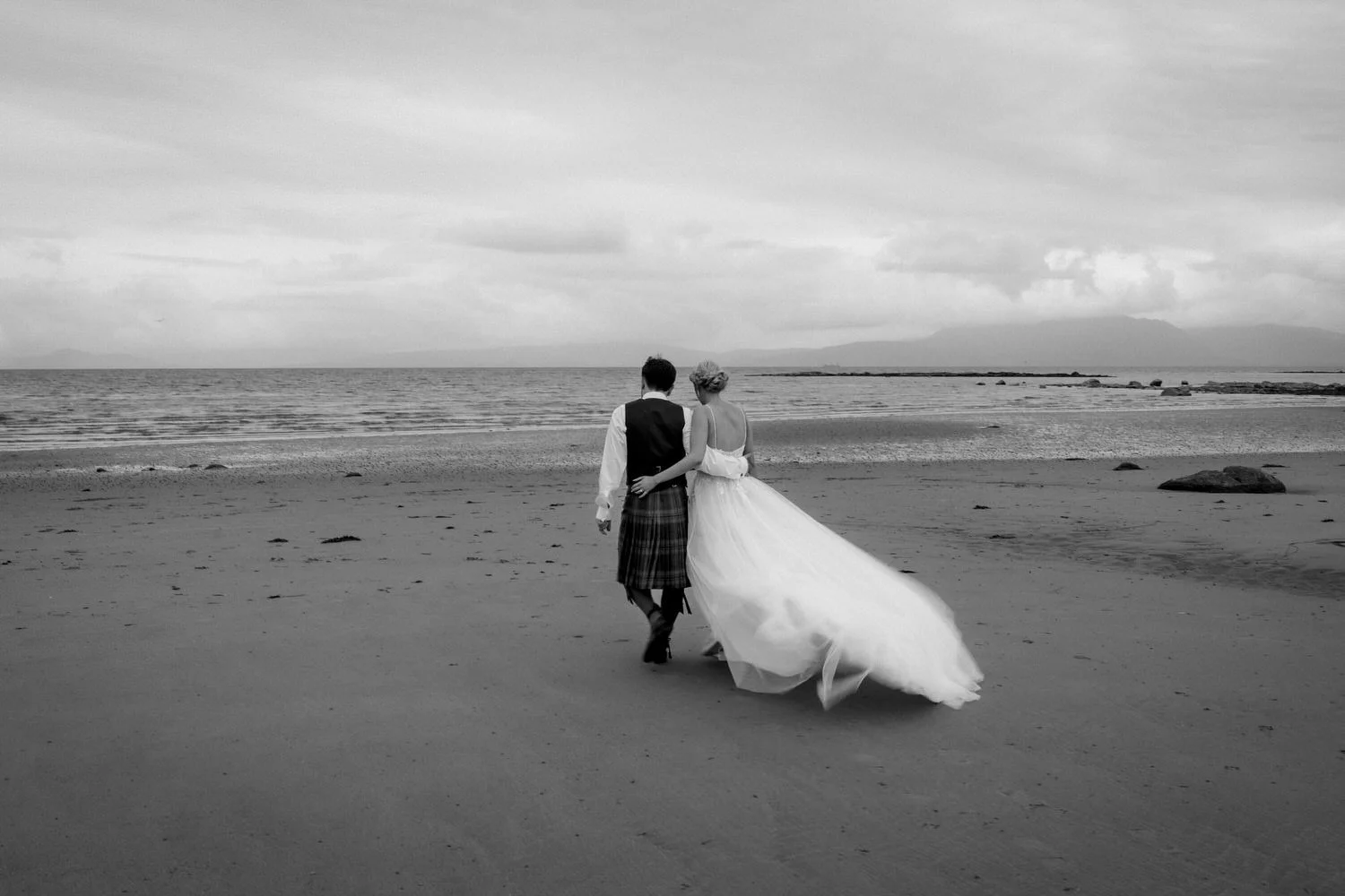 Black and white photo of a bride and groom walking on a beach, holding each other, with the ocean and cloudy sky in the background.