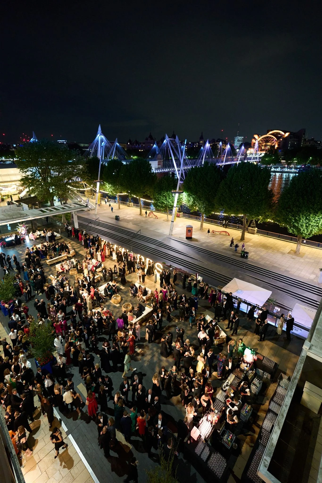 Nighttime view of a crowded outdoor event or party with numerous people, food and drink stalls, and a scenic cityscape with illuminated modern bridge, trees, and buildings in the background.