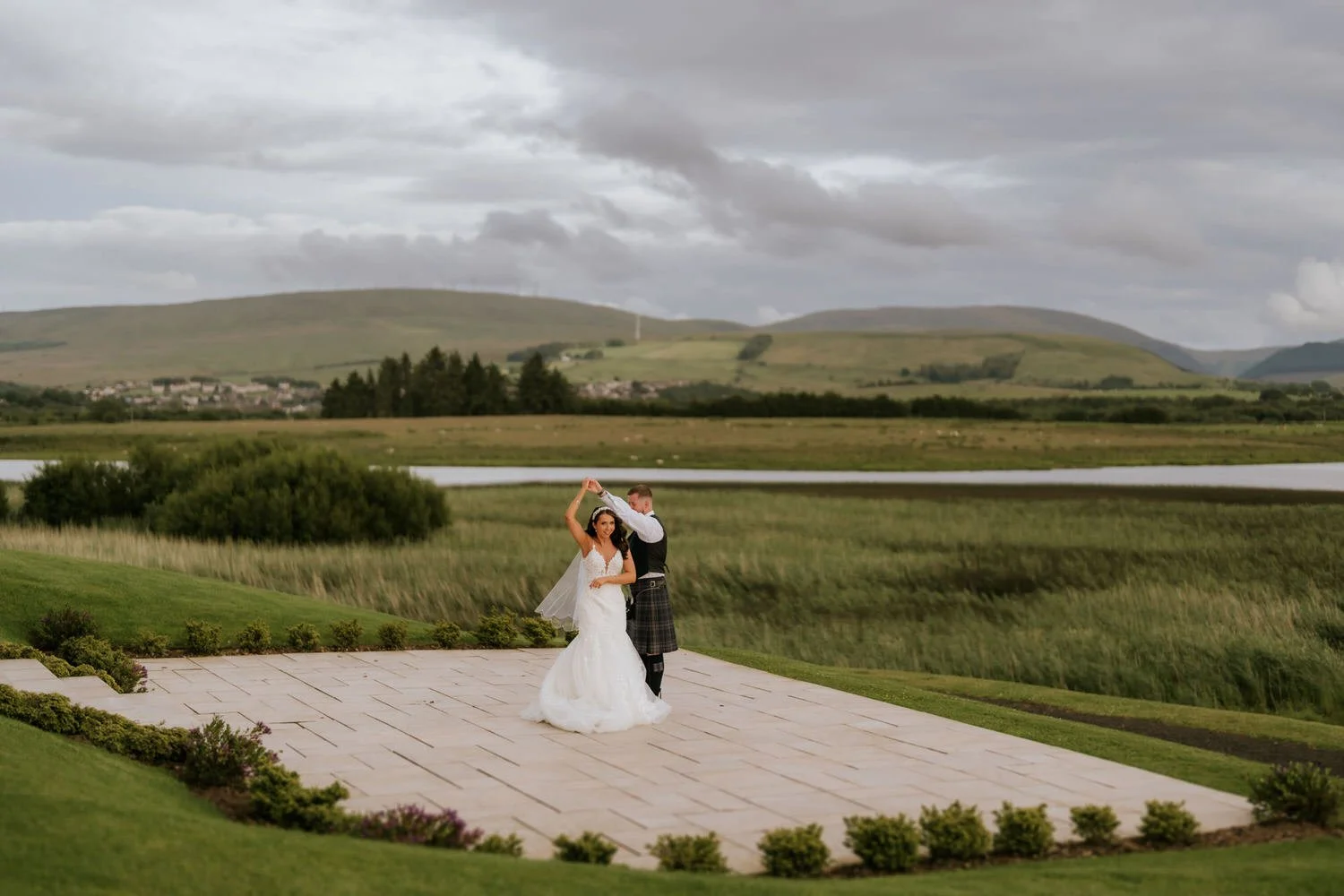 couple dancing on outdoor patio in wedding attire with scenic landscape of hills, water, and cloudy sky in background.