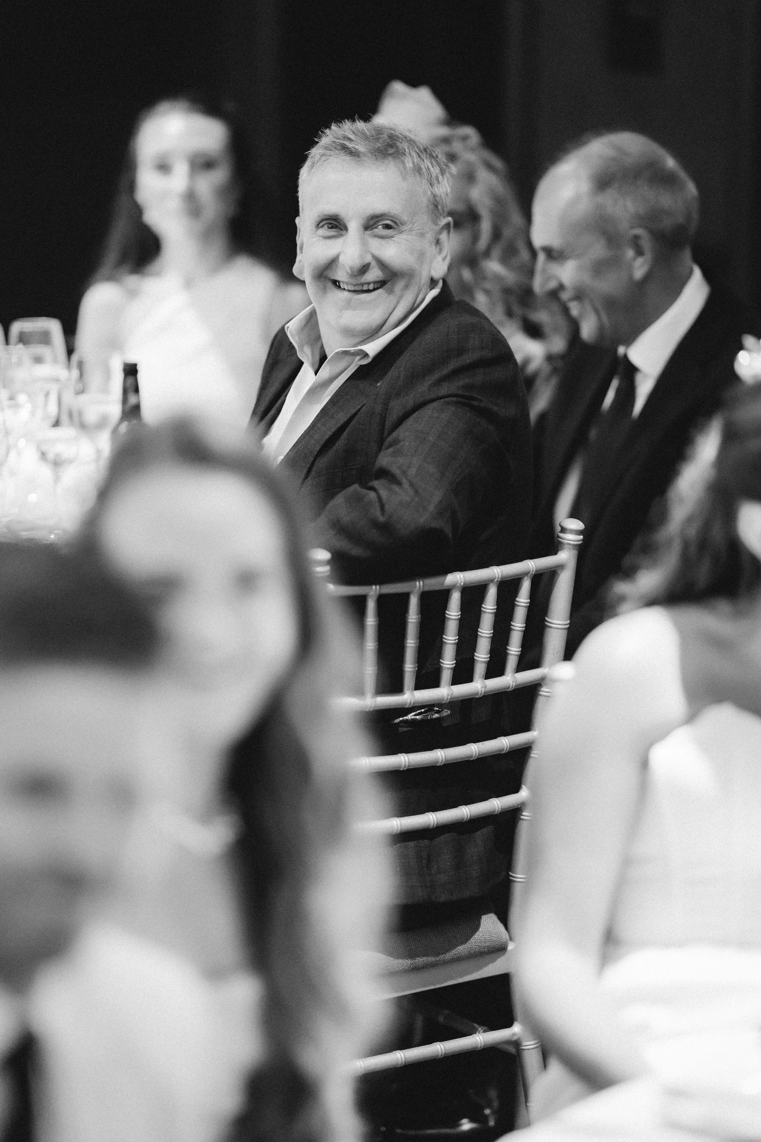 Black-and-white photograph of a guest smiling during the wedding speeches.