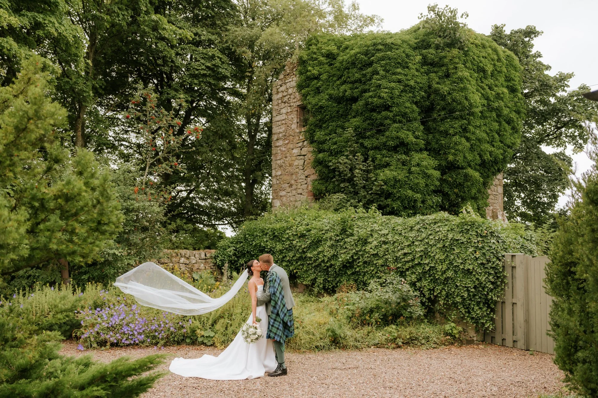 A bride and groom sharing a kiss outdoors, with the bride in a white gown and veil, holding a bouquet, and the groom in a gray suit with a green plaid kilt, standing on a gravel path surrounded by greenery and blooming flowers, with an old stone towe
