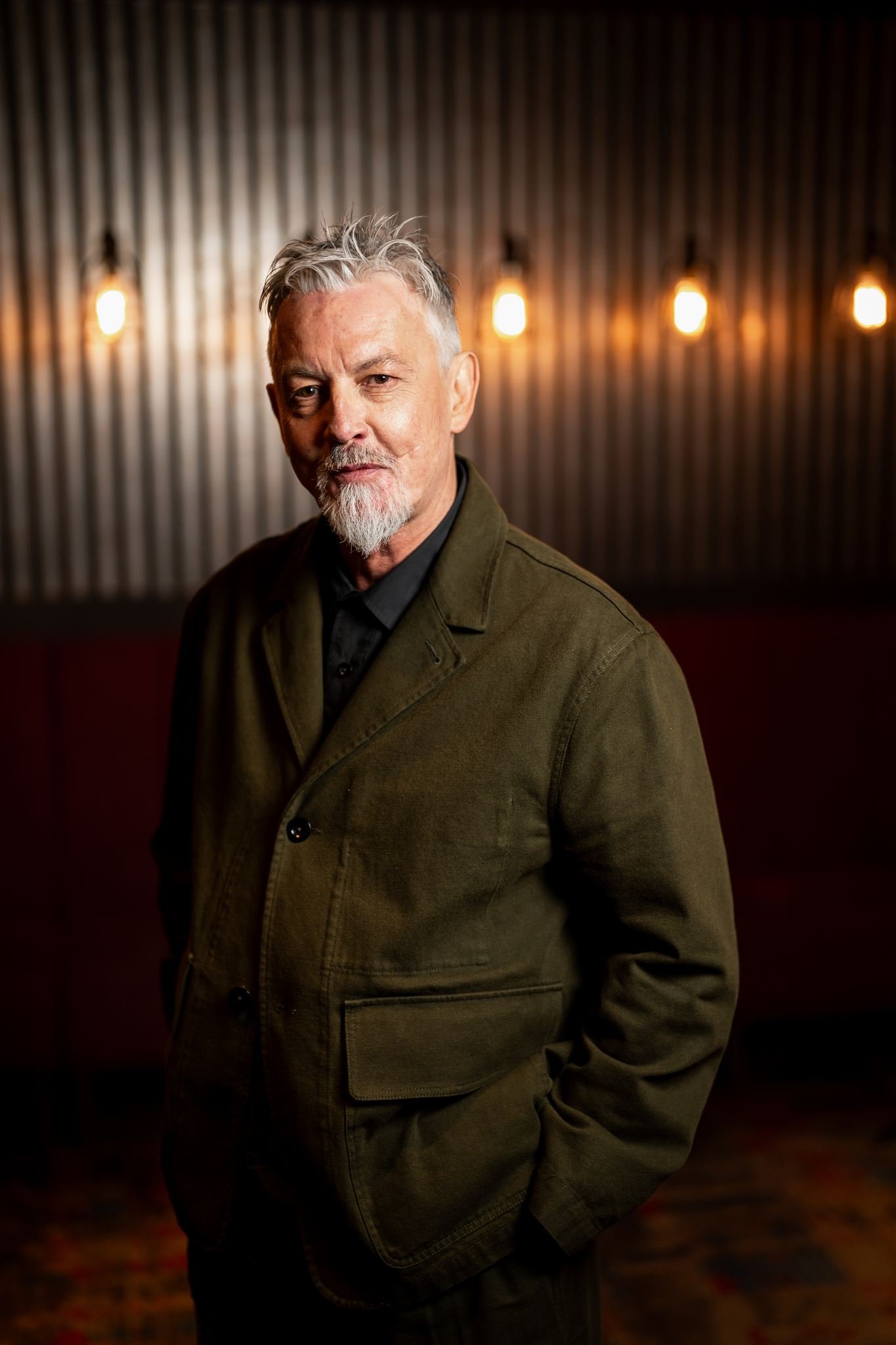 A mature man with gray hair and beard wearing a green jacket, standing in a dimly lit room with a wooden wall and five hanging light bulbs in the background.