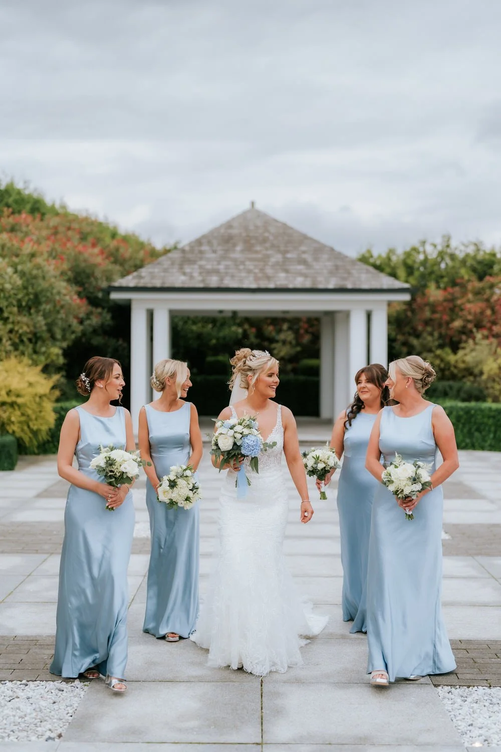 Bride and four bridesmaids walking outdoors, holding bouquets, with greenery and a small gazebo in the background.