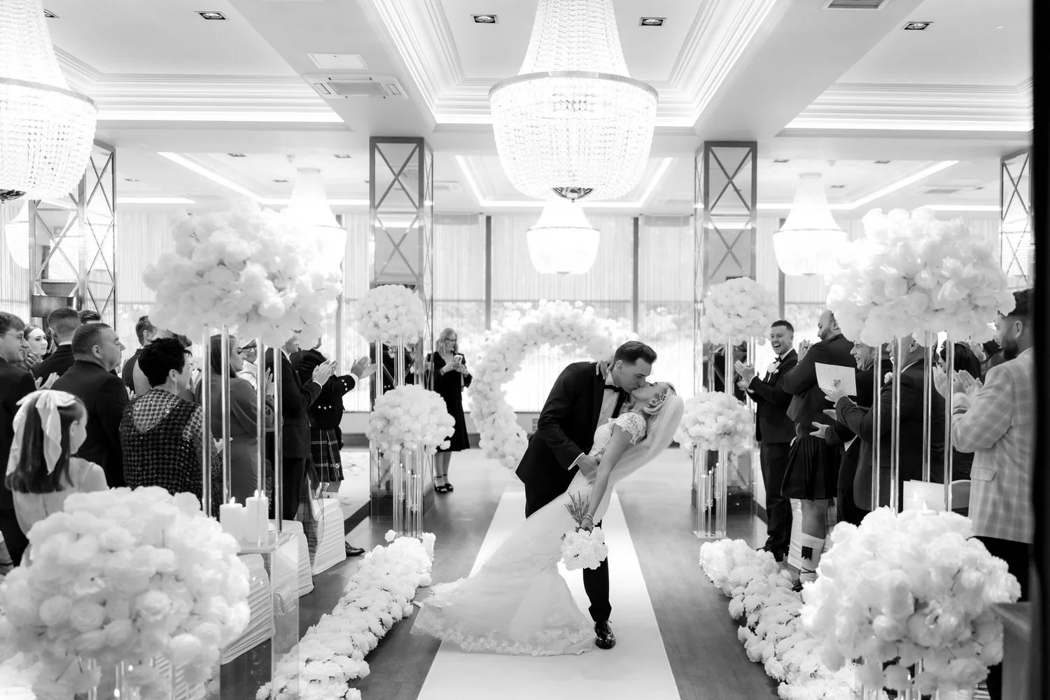 A black and white photo of a bride and groom sharing a kiss during their wedding ceremony in an elegant indoor venue decorated with large floral arrangements, chandeliers, and guests applauding.