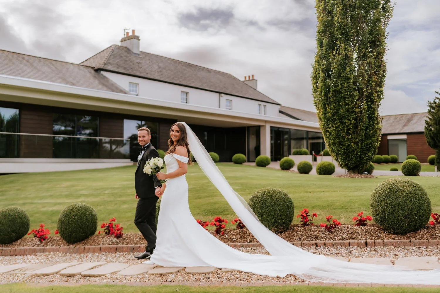 A bride and groom holding hands and walking outside on a wedding day, the bride in a white wedding gown with a long veil and the groom in a black tuxedo, with a modern building and manicured lawn in the background.