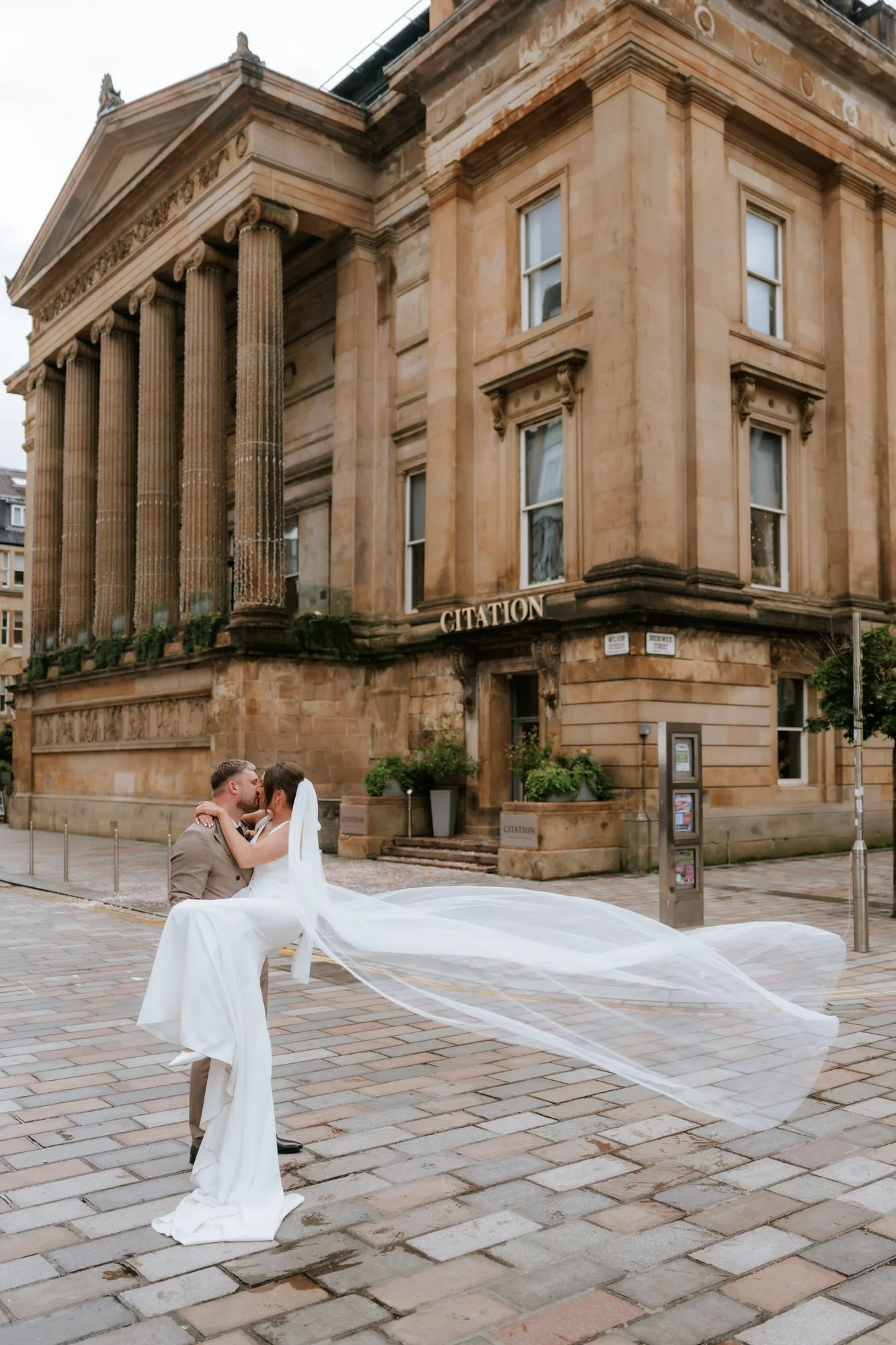 A wedding couple kissing, with the groom lifting the bride, in front of a historic sandstone building labeled 'Citation' with columns and decorative architecture.