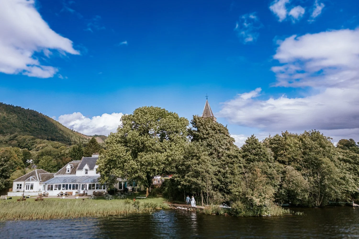 A lakeside scene with a large church surrounded by trees, a white house with a glass extension, on a bright day with a few clouds in the sky.