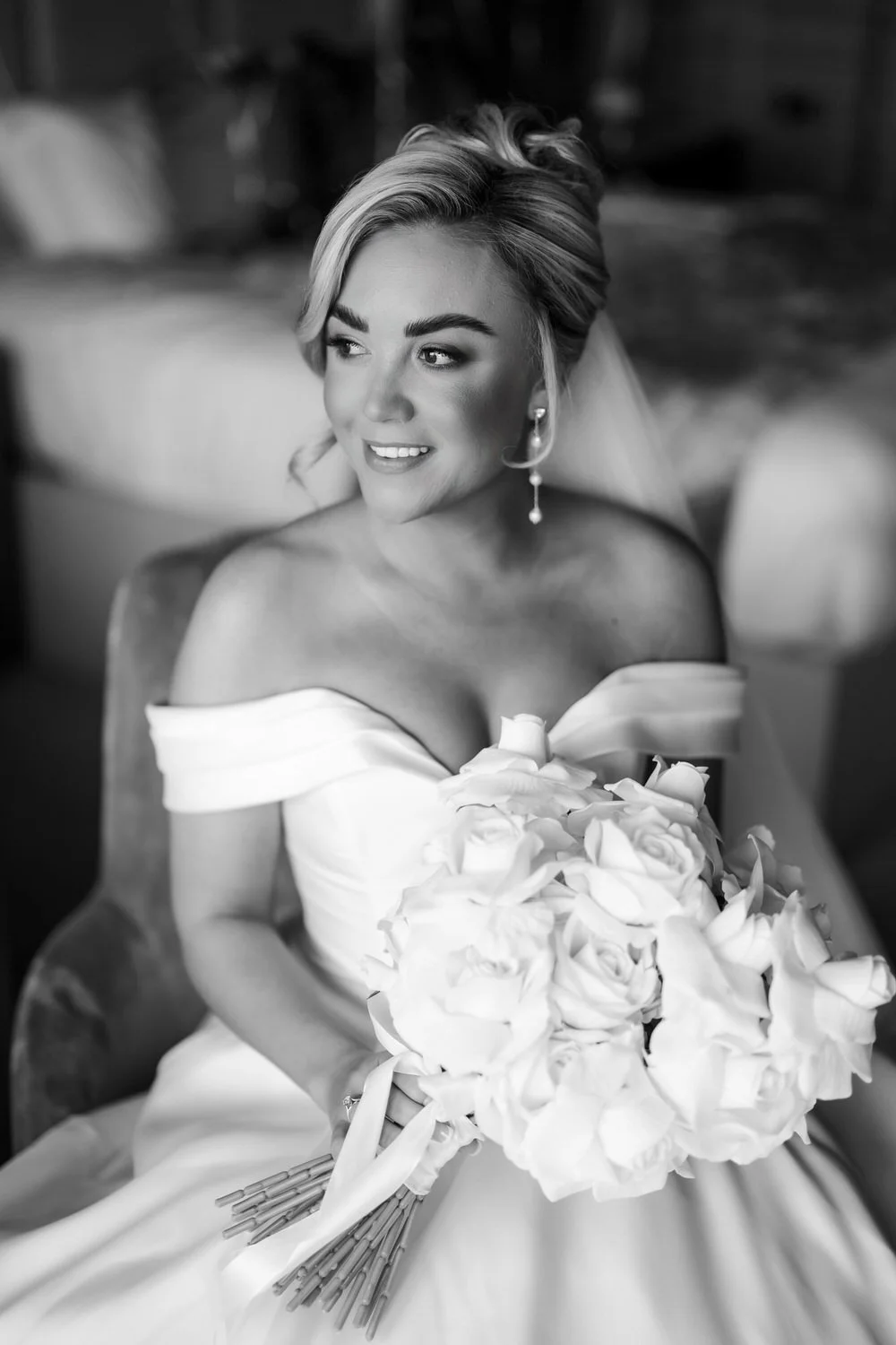 A black-and-white photo of a smiling bride holding a bouquet of white flowers, wearing an off-shoulder wedding dress, with her hair styled and earrings.
