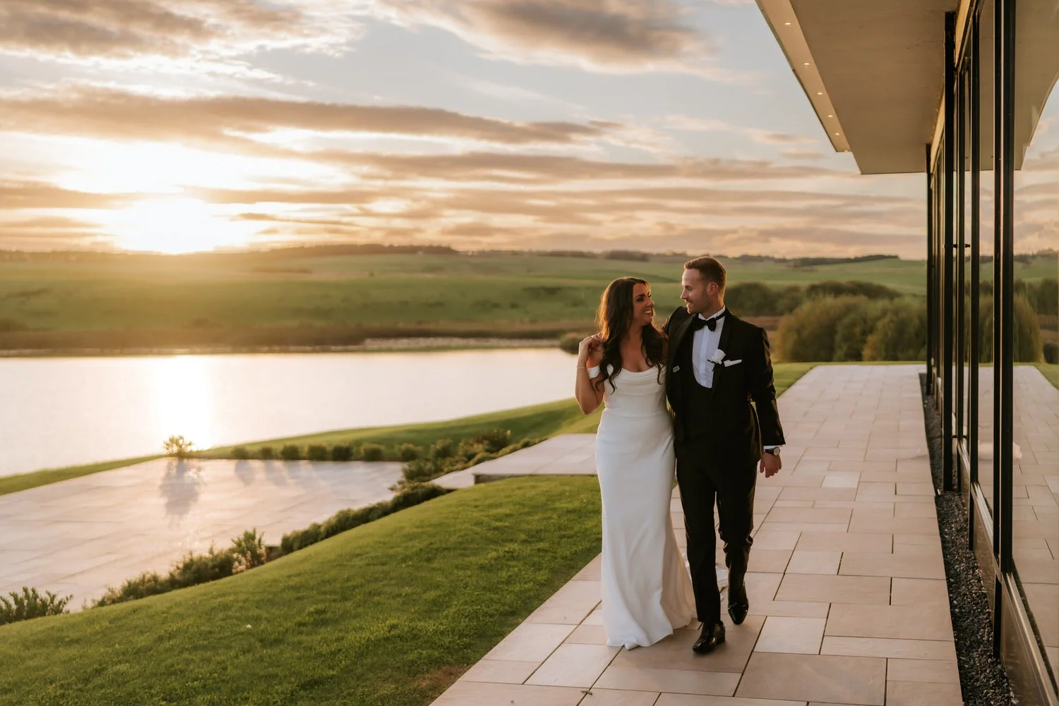 A bride in a white wedding dress and a groom in a black tuxedo walking arm in arm on a balcony during sunset, overlooking a lake and green fields.