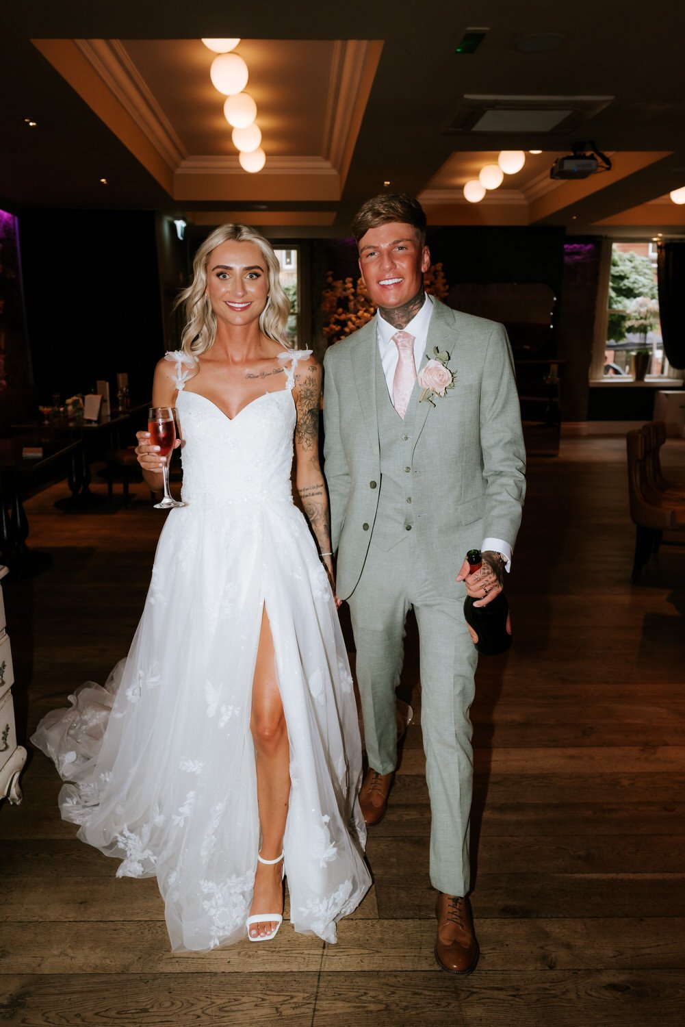 A bride and groom standing together indoors at their wedding reception, the bride holding a glass of wine and the groom holding a bottle of champagne, both smiling at the camera.