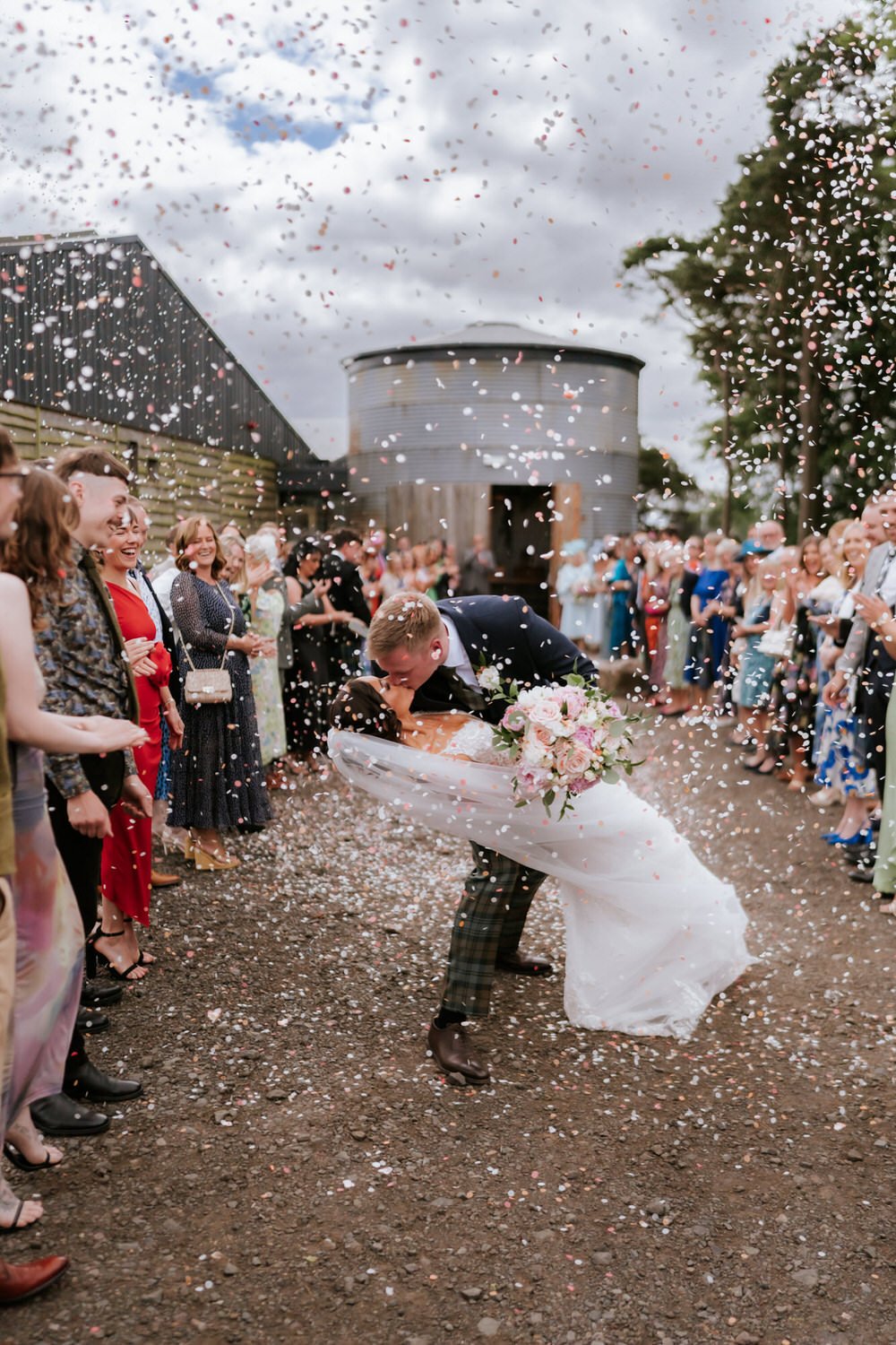 A joyful bride and groom sharing a kiss while the groom dips the bride in front of a crowd during their wedding celebration, with confetti falling from the sky.