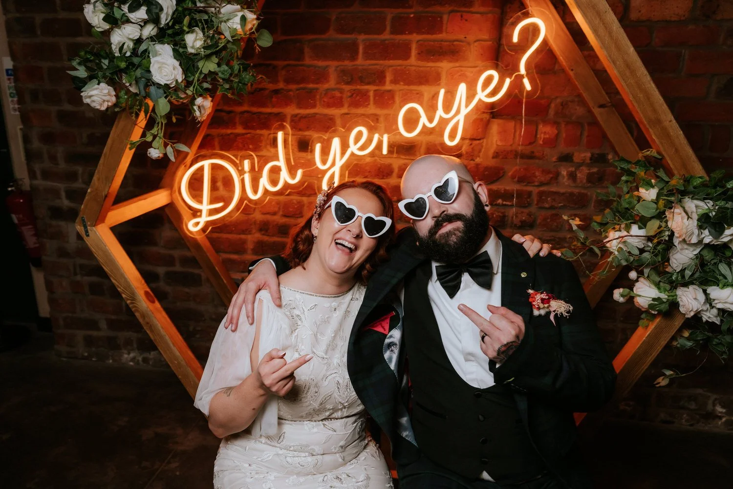 A couple at a celebration with neon sign that reads 'Did yе, aye?' in front of a brick wall, both wearing white heart-shaped sunglasses and making playful gestures, surrounded by flowers and greenery.