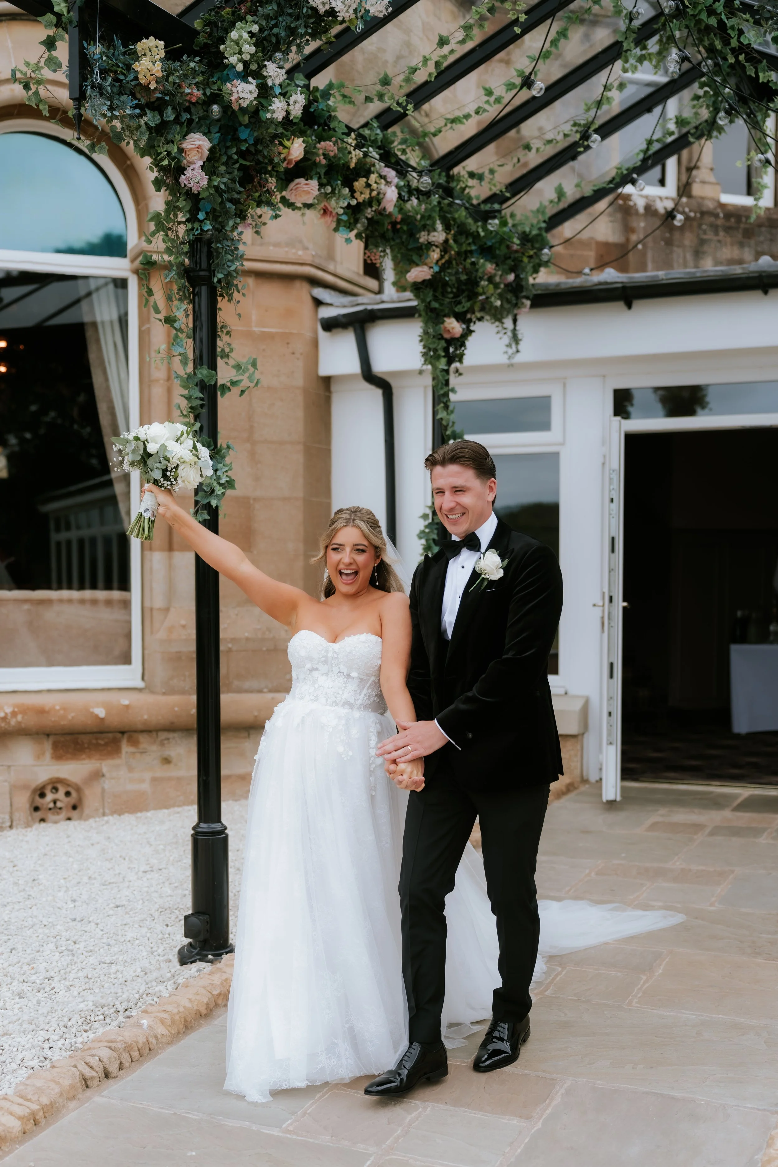 Emma raising her bouquet beside Frazer outside the pavilion entrance.
