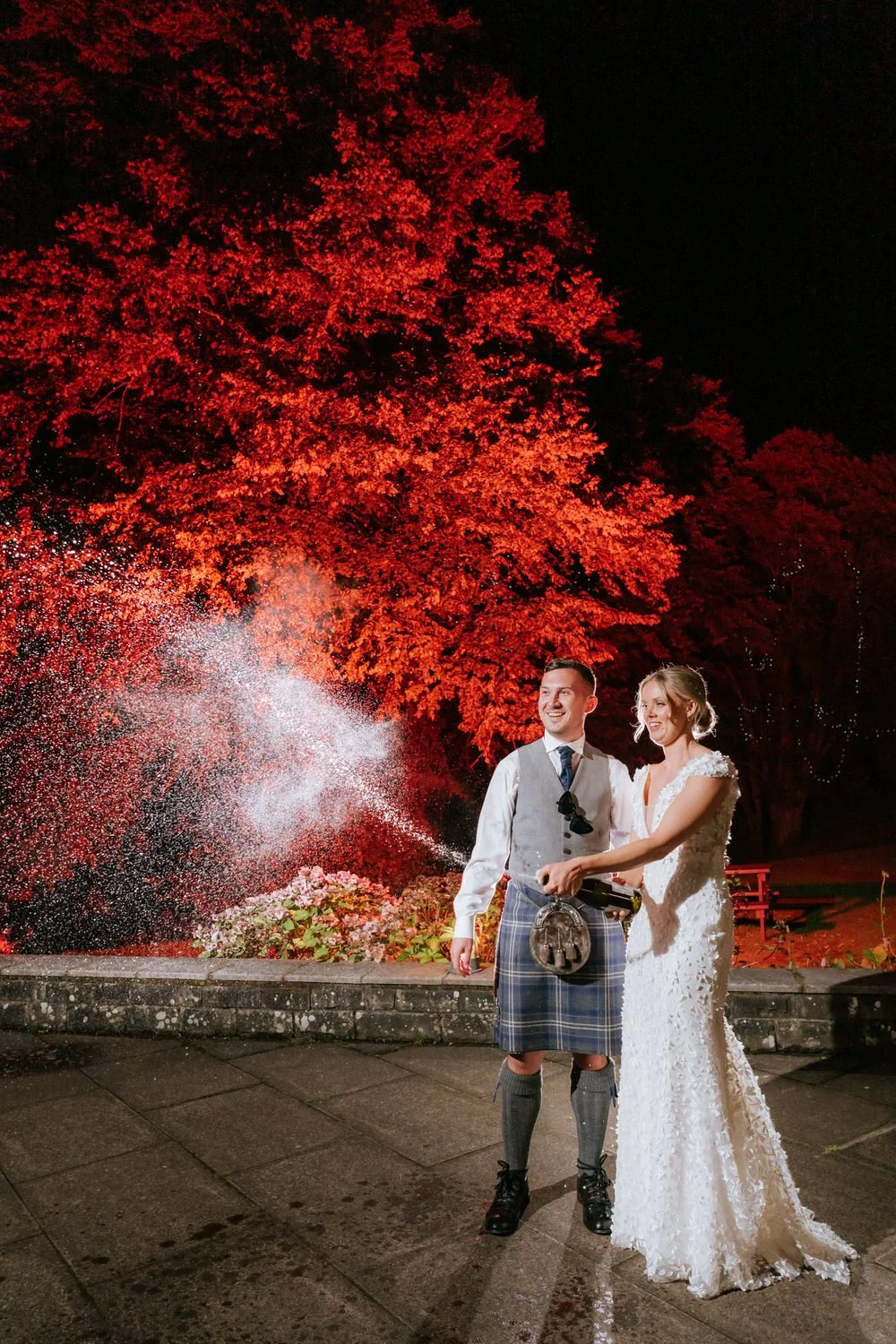 Couple celebrating a wedding outdoors at night, with a large red-leaved tree in the background, and spraying champagne.