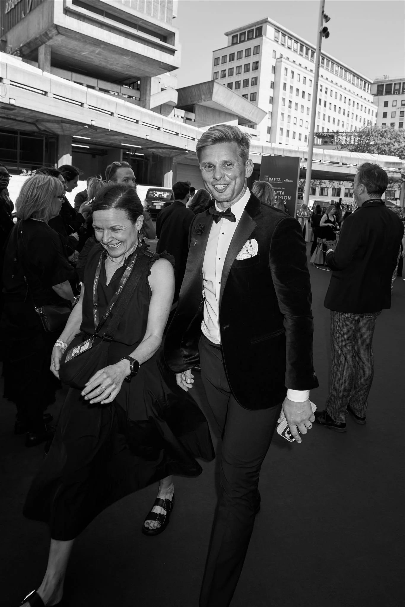 A black and white photo of two people, a woman and a man, dressed in formal attire, smiling and walking together outdoors at an event, with other people and tall buildings in the background.