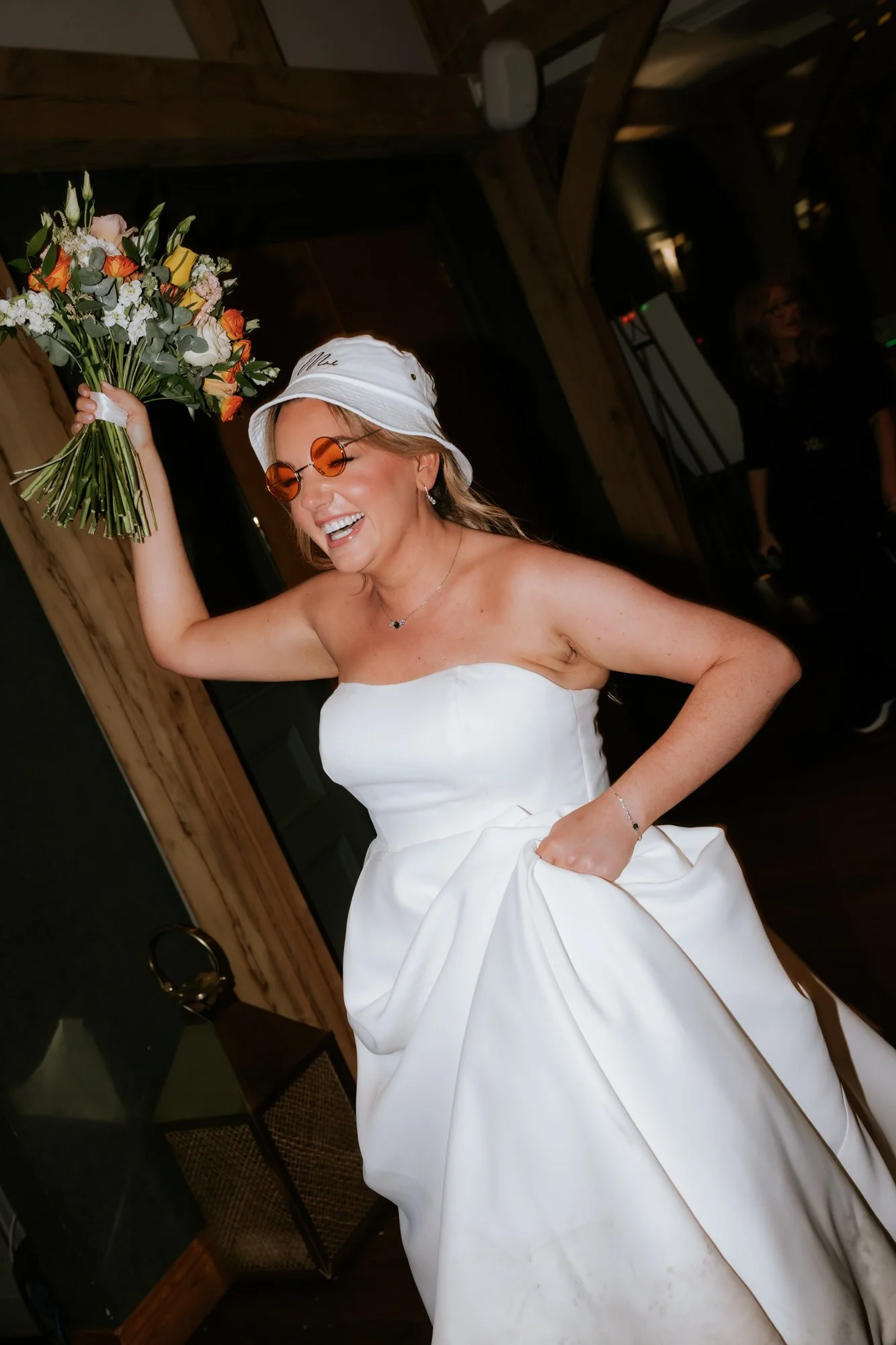 A woman in a white wedding dress, wearing a white hat and orange sunglasses, holding a bouquet of flowers, and smiling happily.