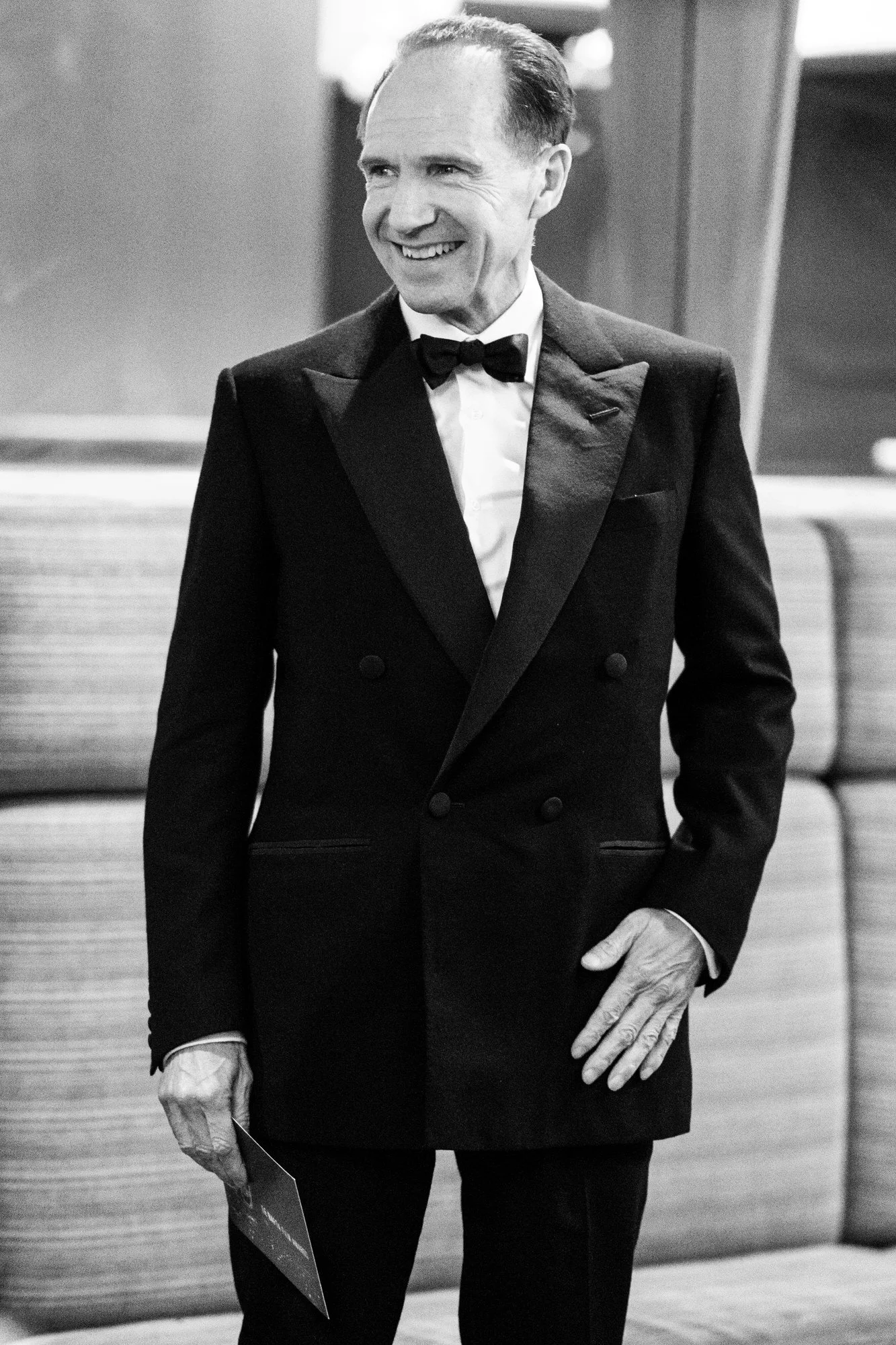 Ralph Fiennes smiling backstage at the 2025 BAFTA Film Awards, in black and white.