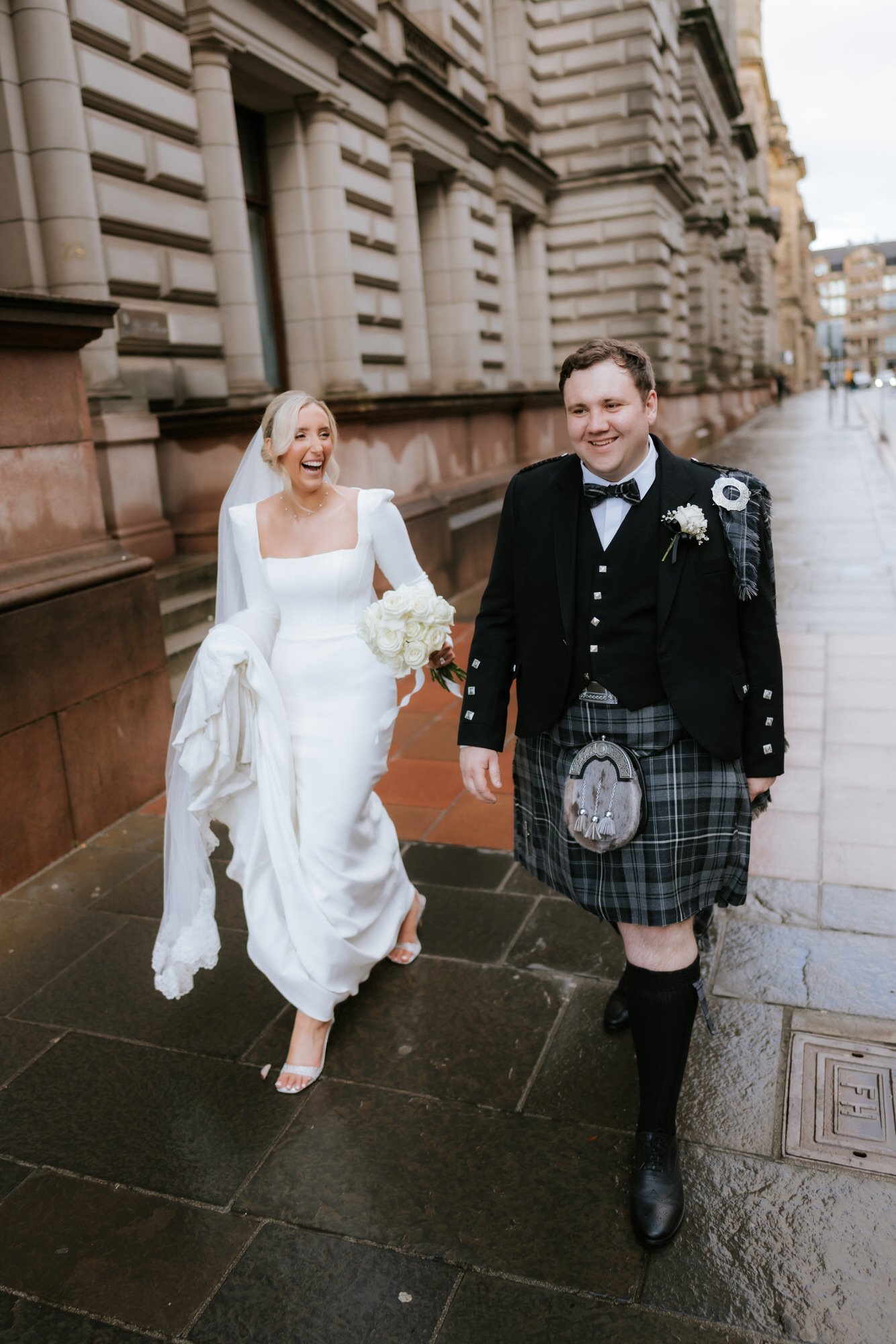 A bride and groom walking on a city street, smiling and laughing. The bride is wearing a white wedding gown and holding a bouquet of white roses. The groom is dressed in traditional Scottish attire, including a kilt and jacket.