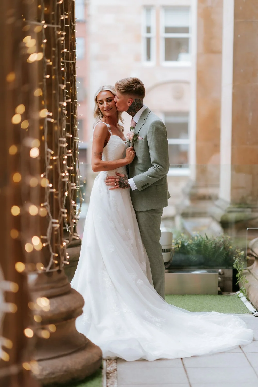 A bride and groom share a kiss in a wedding photo, with fairy lights and a building facade in the background.