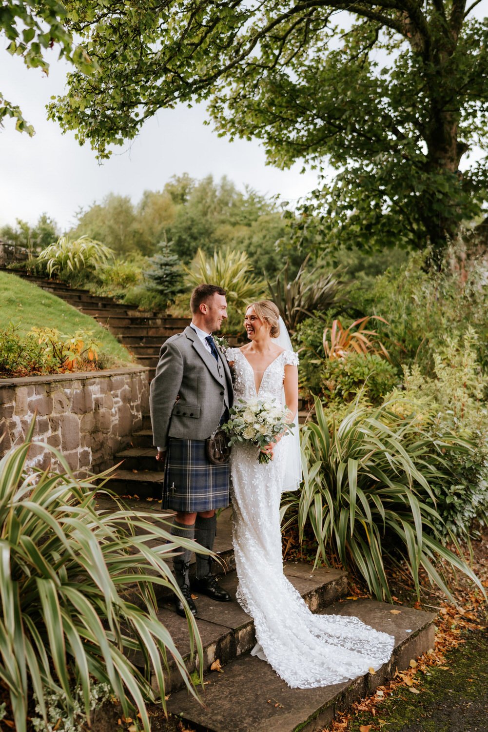 A bride and groom standing on stone steps in a garden, smiling at each other. The bride wears a white lace wedding gown with a bouquet, and the groom wears traditional Scottish attire including a kilt.