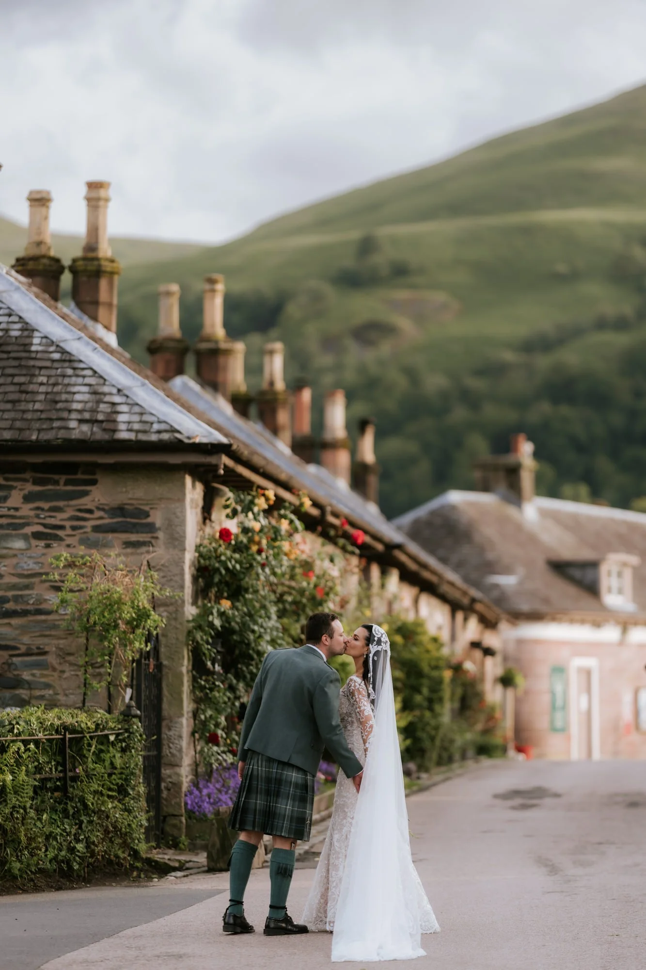 A bride and groom kissing on a quiet street in a village with stone houses, lush greenery, and hills in the background.
