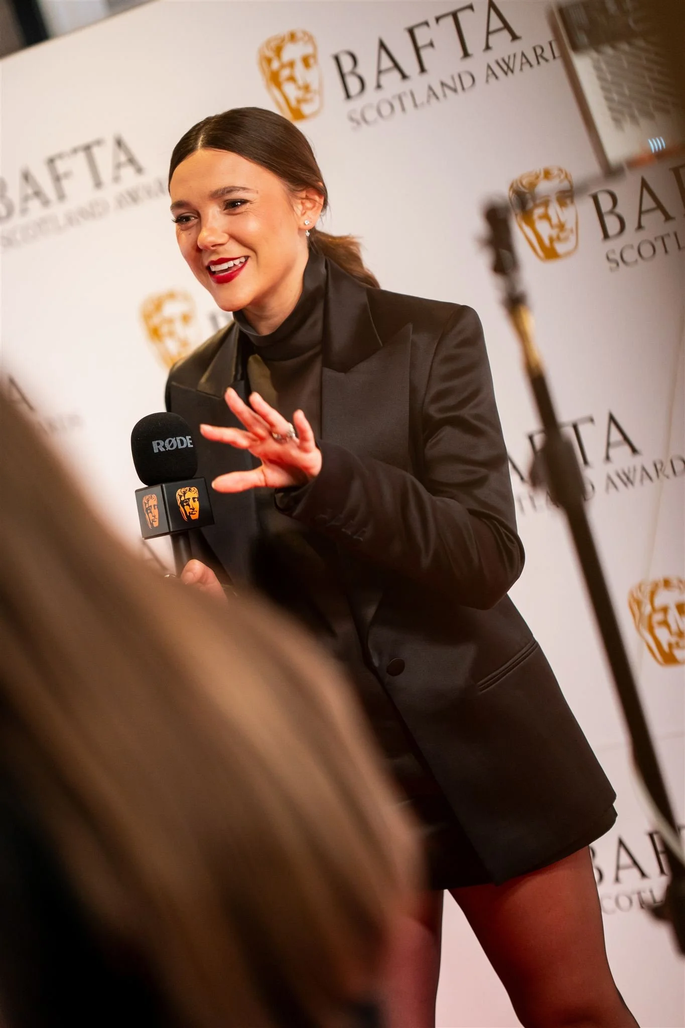 A woman with dark brown hair in a ponytail, wearing a black blazer and a dark top, speaking into a microphone at the BAFTA Scotland Awards event, with a BAFTA Scotland backdrop behind her.