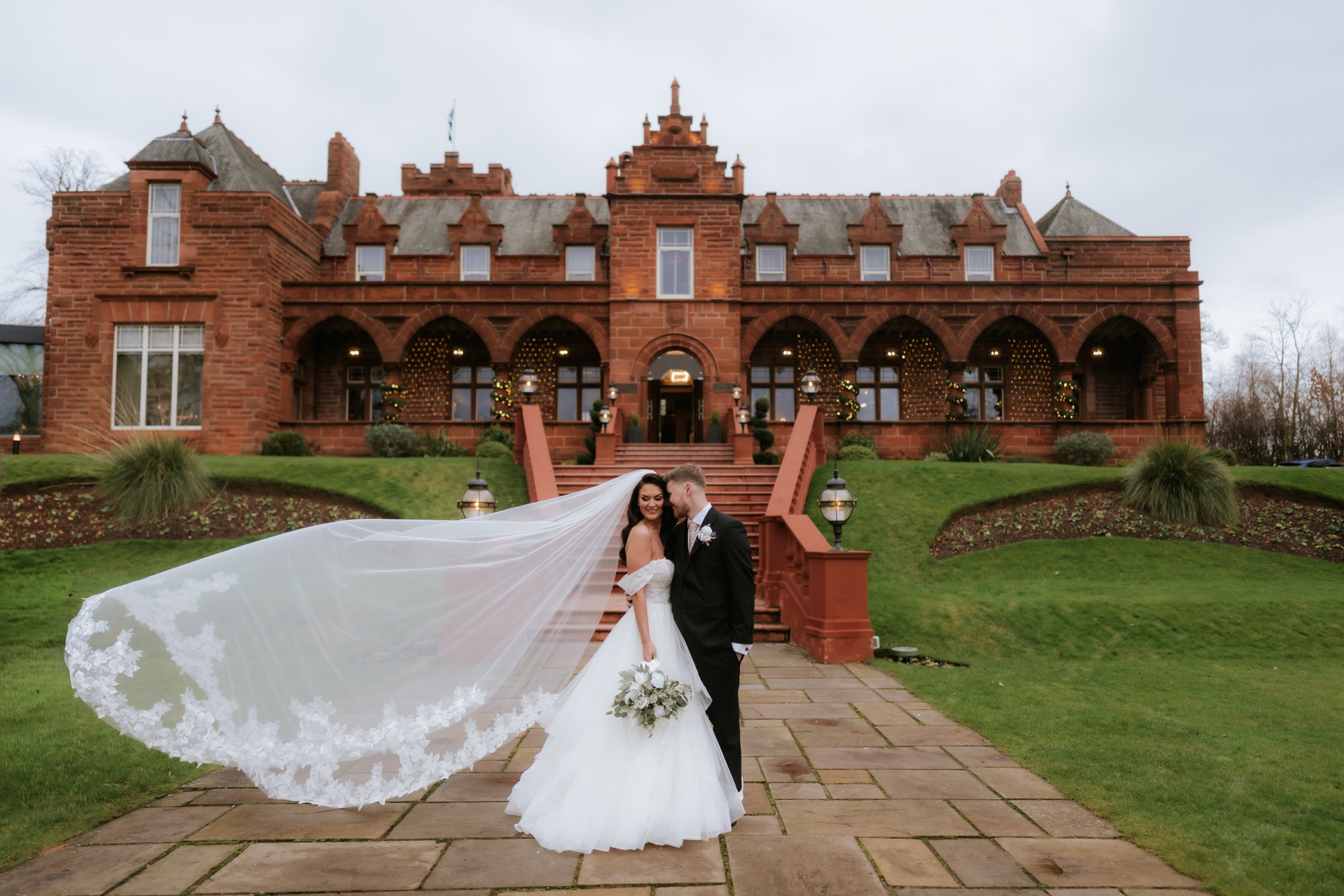 A bride and groom in wedding attire standing on a stone path in front of a historic red brick mansion, with the bride’s long veil flowing behind her.
