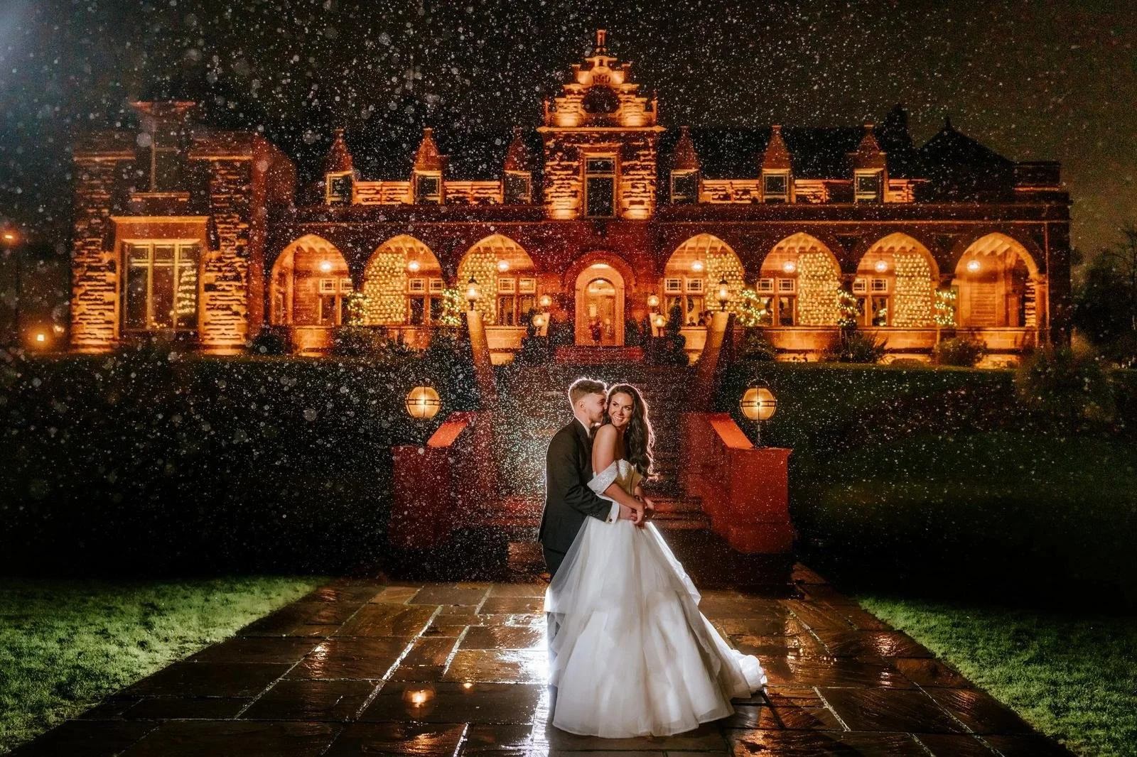 Bride and groom outside illuminated Boclair House at night, with rain and reflections on the ground.