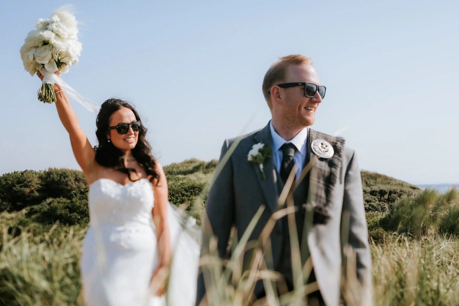 A newlywed couple walking outdoors in a grassy field on a sunny day, with the bride holding a bouquet in the air and smiling, and the groom smiling and looking away, both wearing sunglasses.