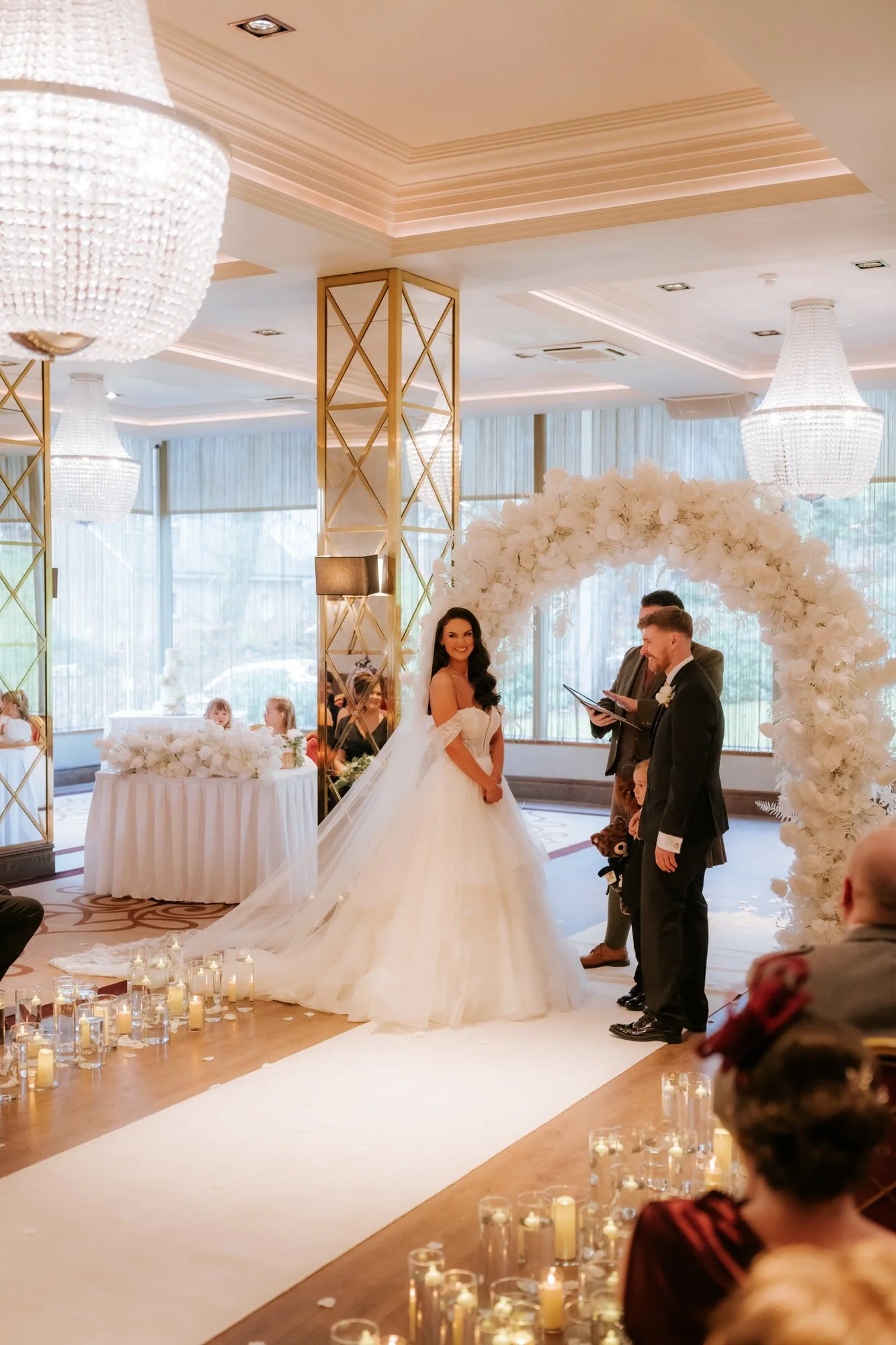 Bride smiling during the ceremony beneath a white floral arch
