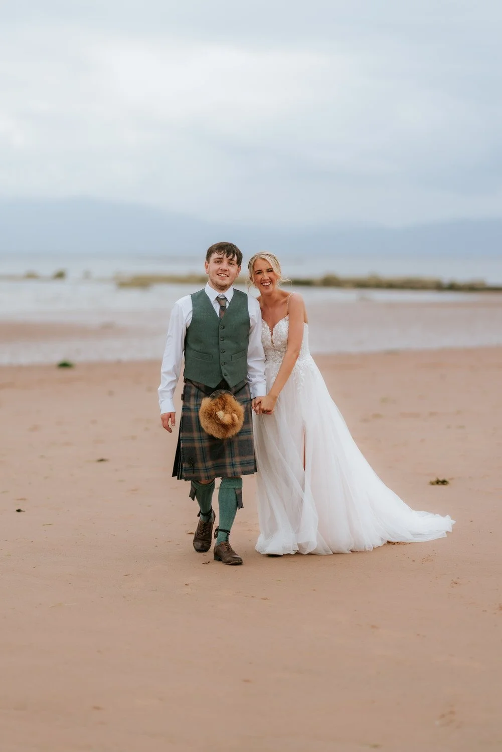 A couple dressed in wedding attire walking on a beach, with the groom in traditional Scottish kilt and the bride in a white wedding dress, holding hands and smiling.