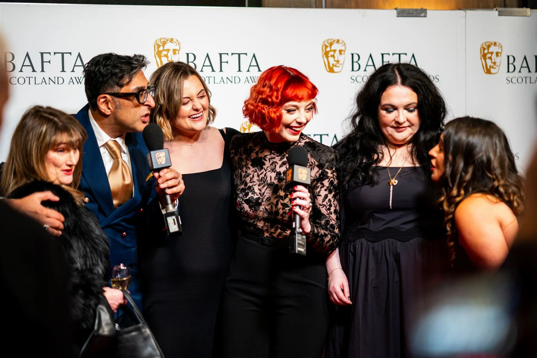 Group of six people at an awards event, standing in front of a backdrop with 'BAFTA Scotland Awards' text and BAFTA logo, engaged in a conversation.