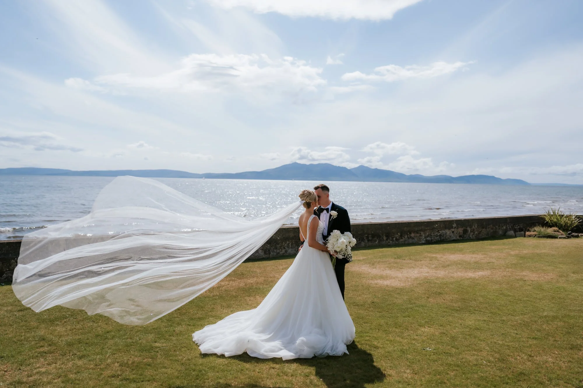 A bride and groom kiss outdoors by the water on their wedding day, with the bride's veil flowing in the breeze and a mountain visible in the background.