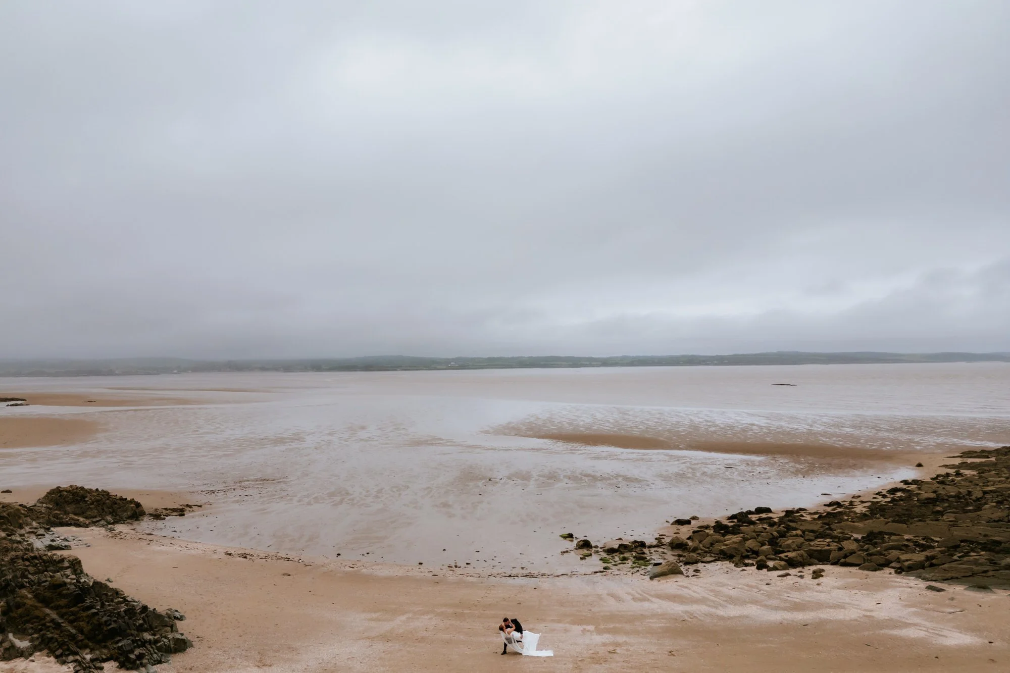A bride and groom embrace on a sandy beach under an overcast sky, with rocks along the shoreline and a distant water view.